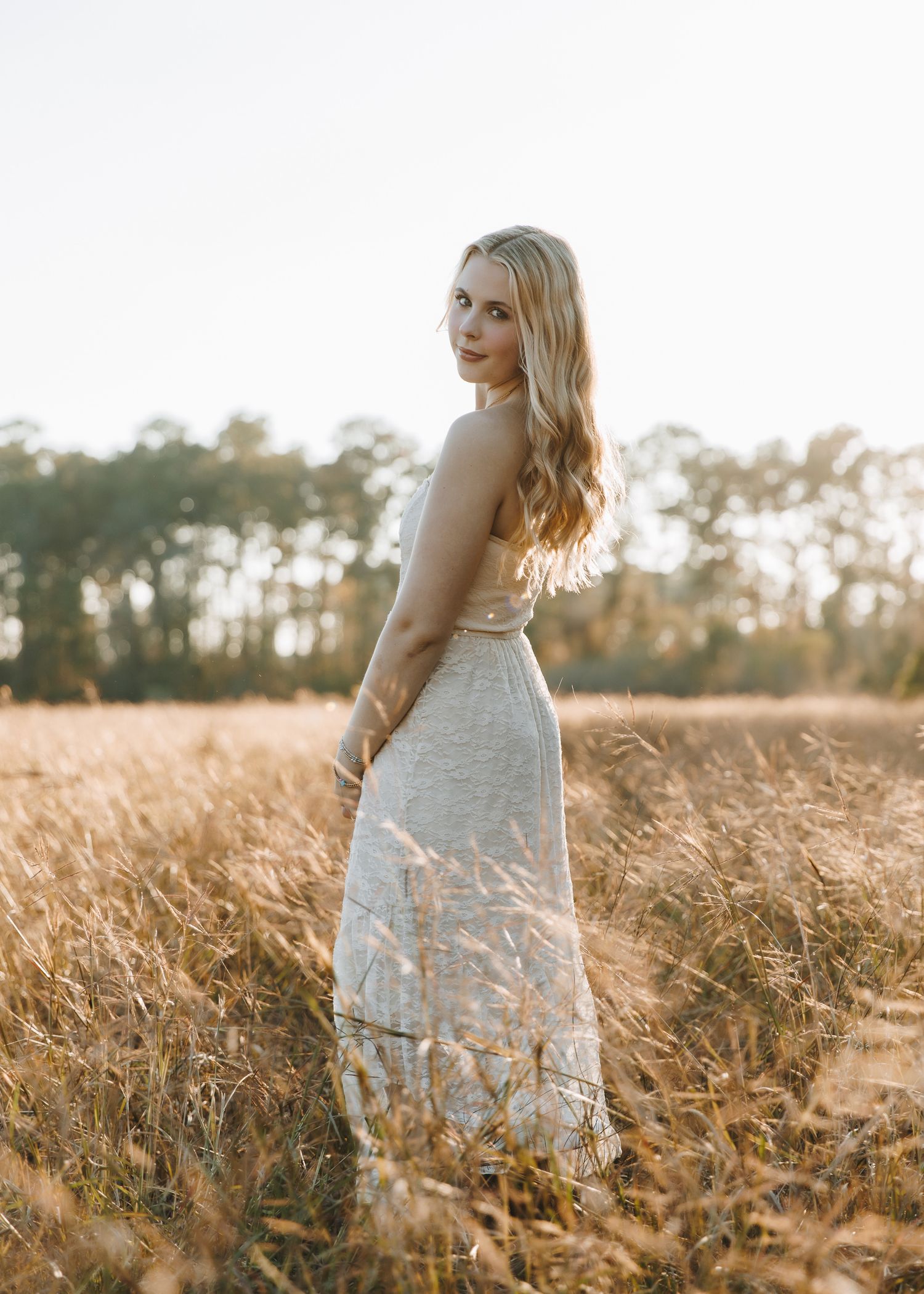 Blonde woman in white maxi dress standing in golden wheat field during golden hour.
