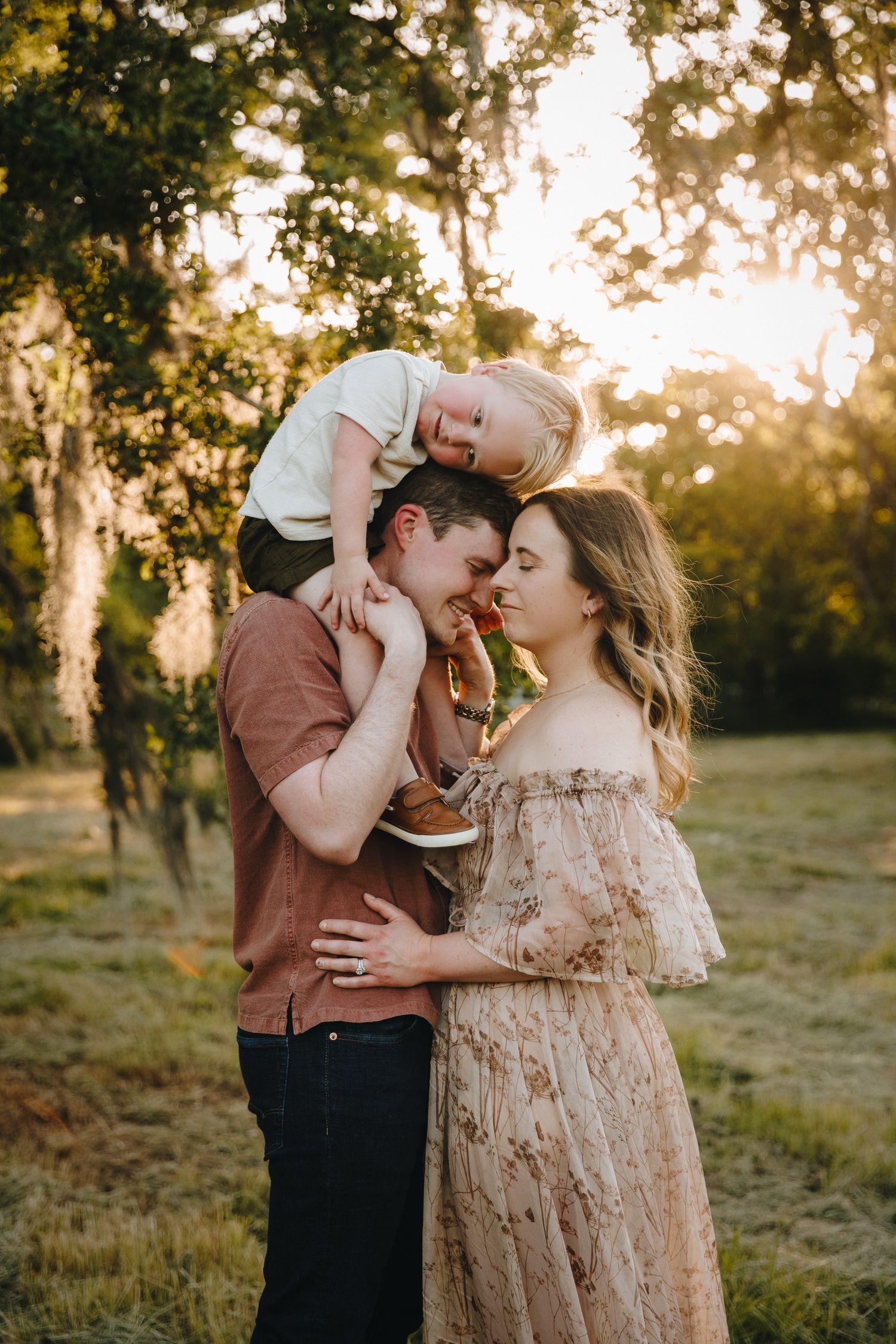 Young family at golden hour, toddler sitting on fathers shoulders while parents share a tender moment.