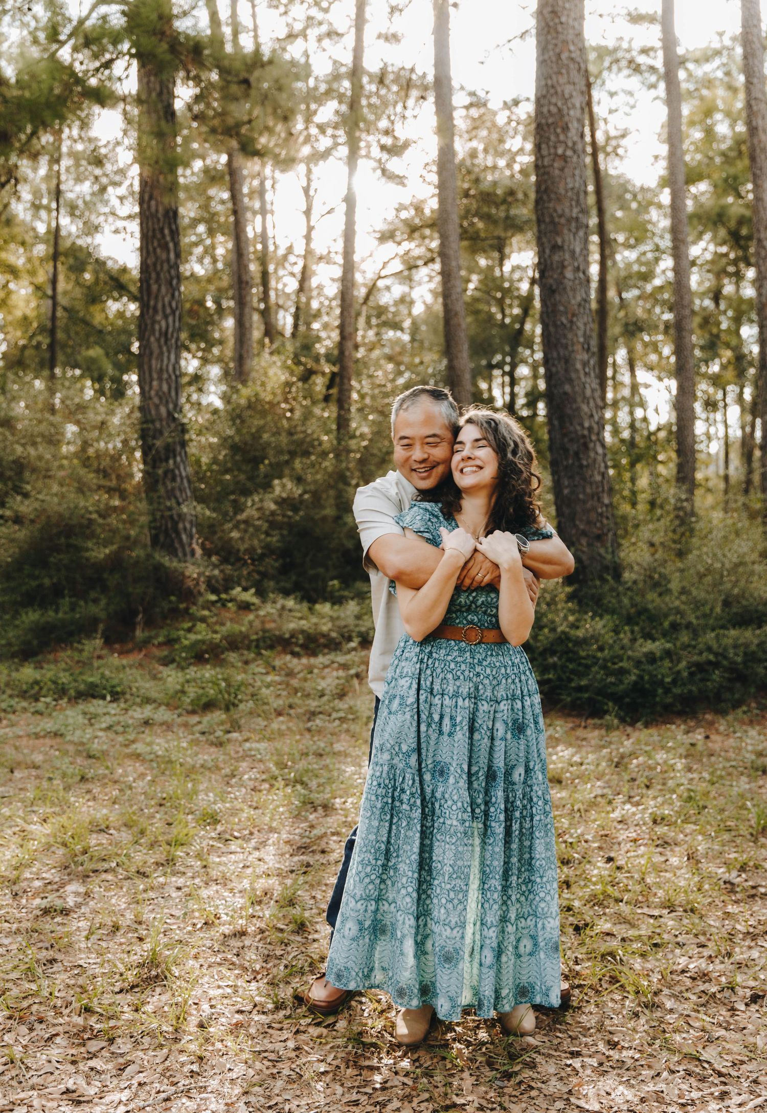 Happy couple embracing and laughing in a pine forest, woman in bohemian teal maxi dress.