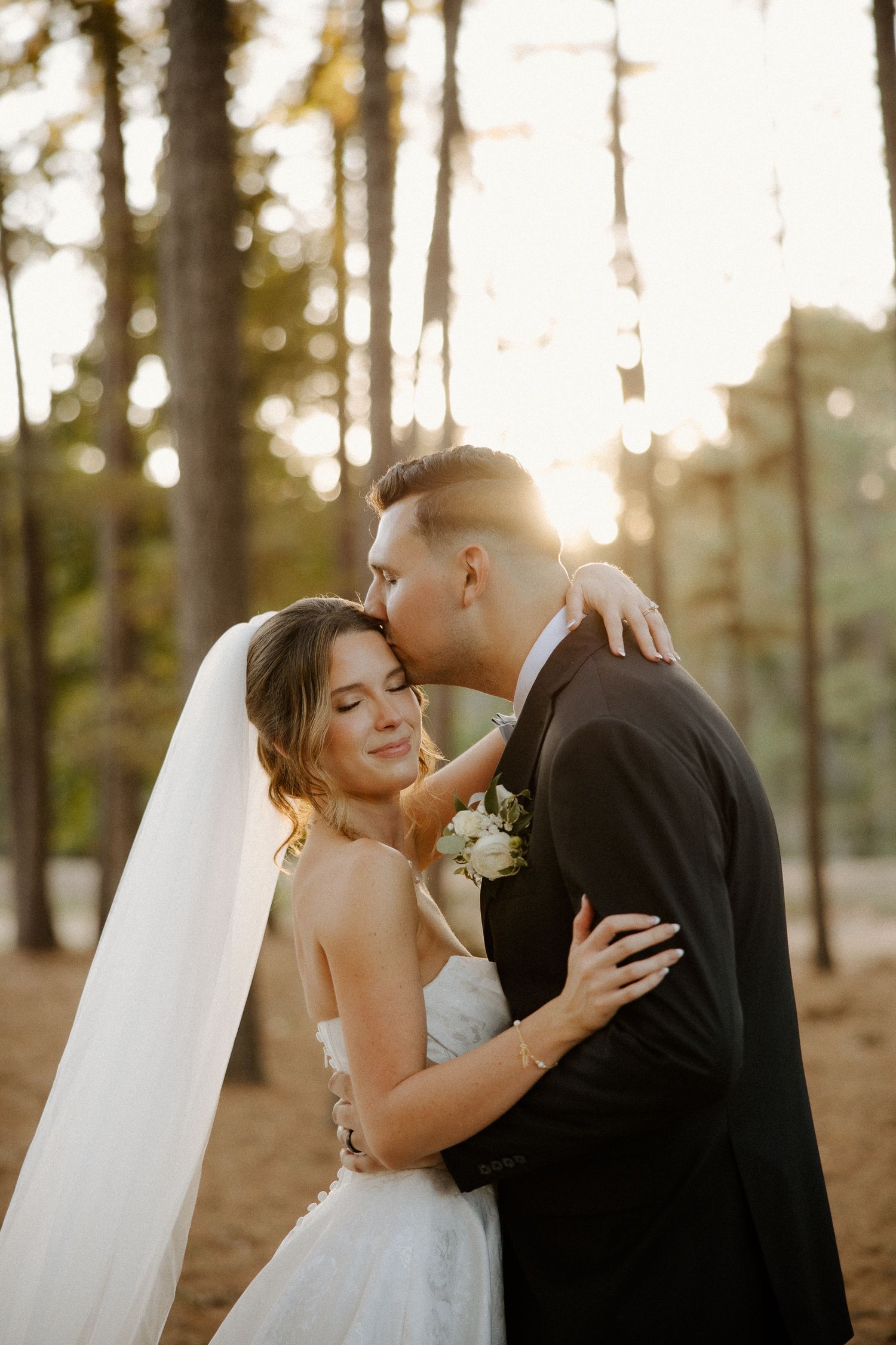 Groom kissing bride on forehead during golden hour portrait in pine forest.