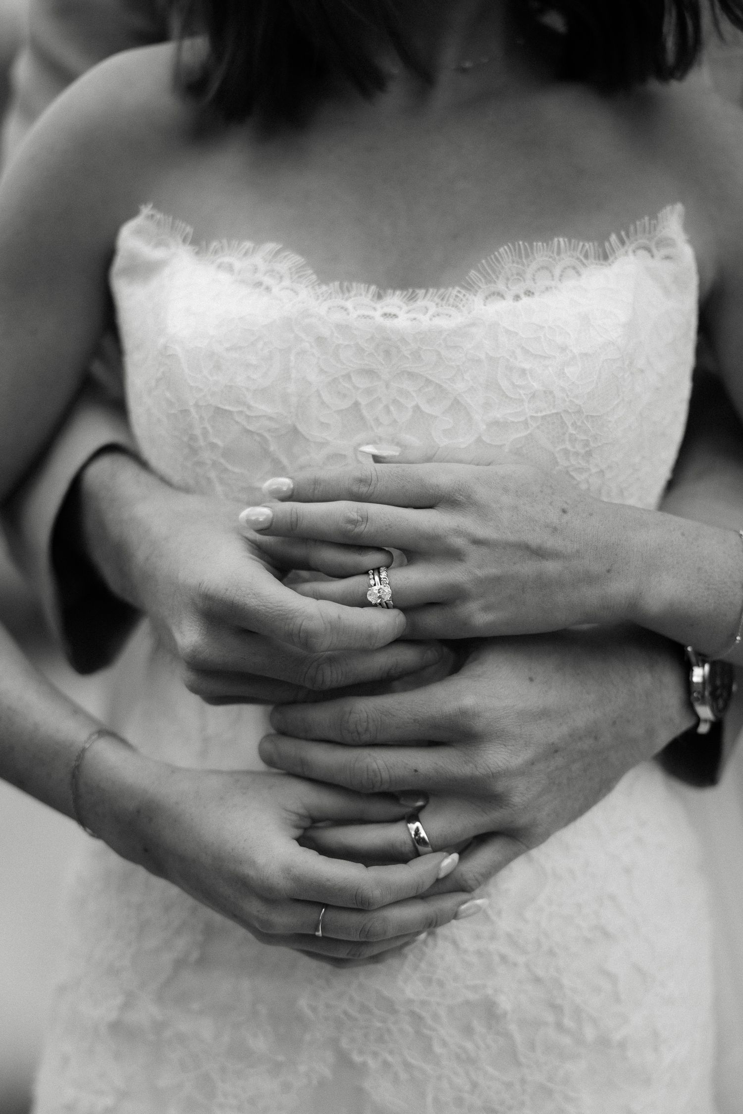 Black and white photo of bride in lace dress with groom's arms wrapped around her, both wearing wedding rings.