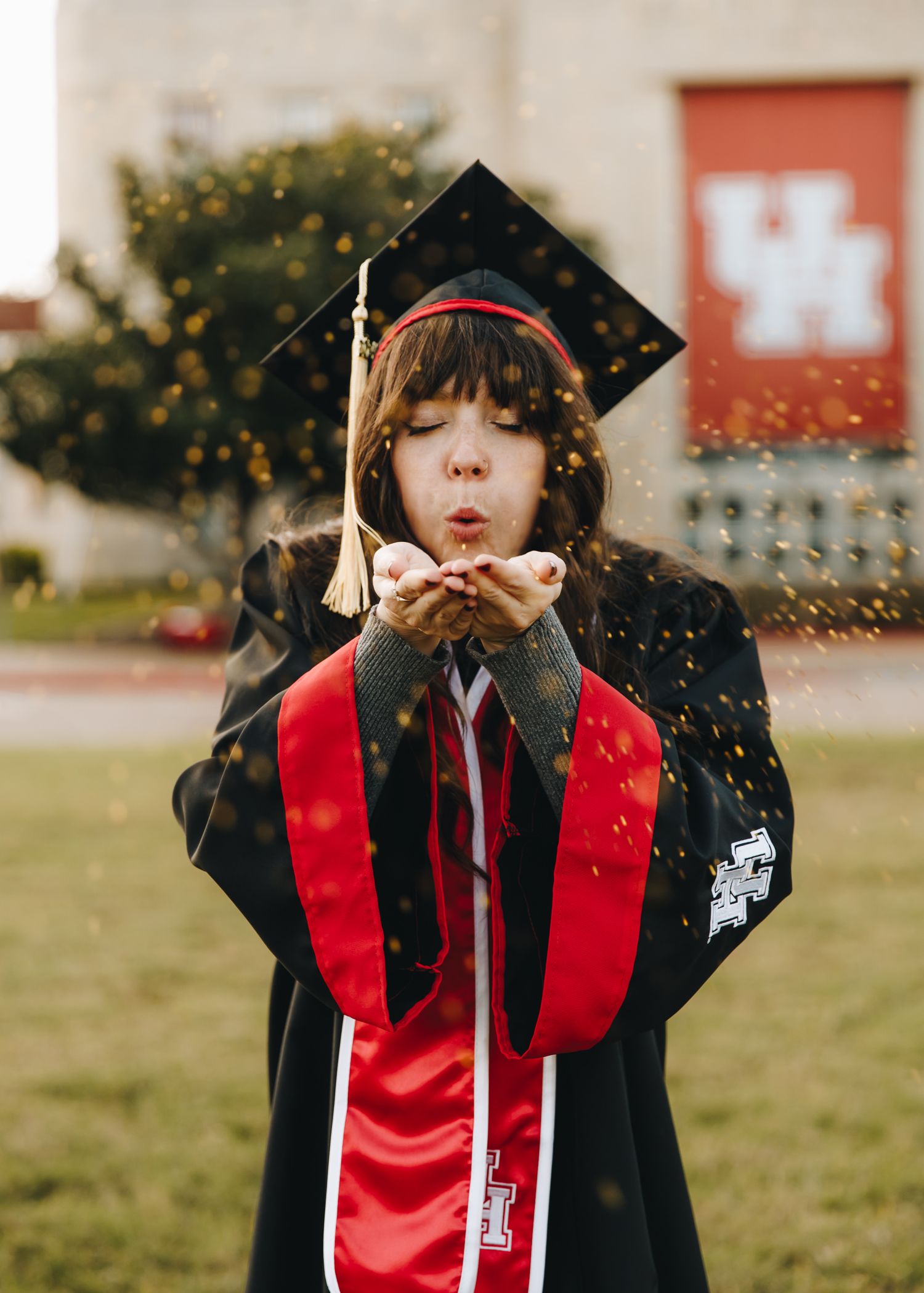 Graduate in UH cap and gown blows gold glitter from hands outside campus building.