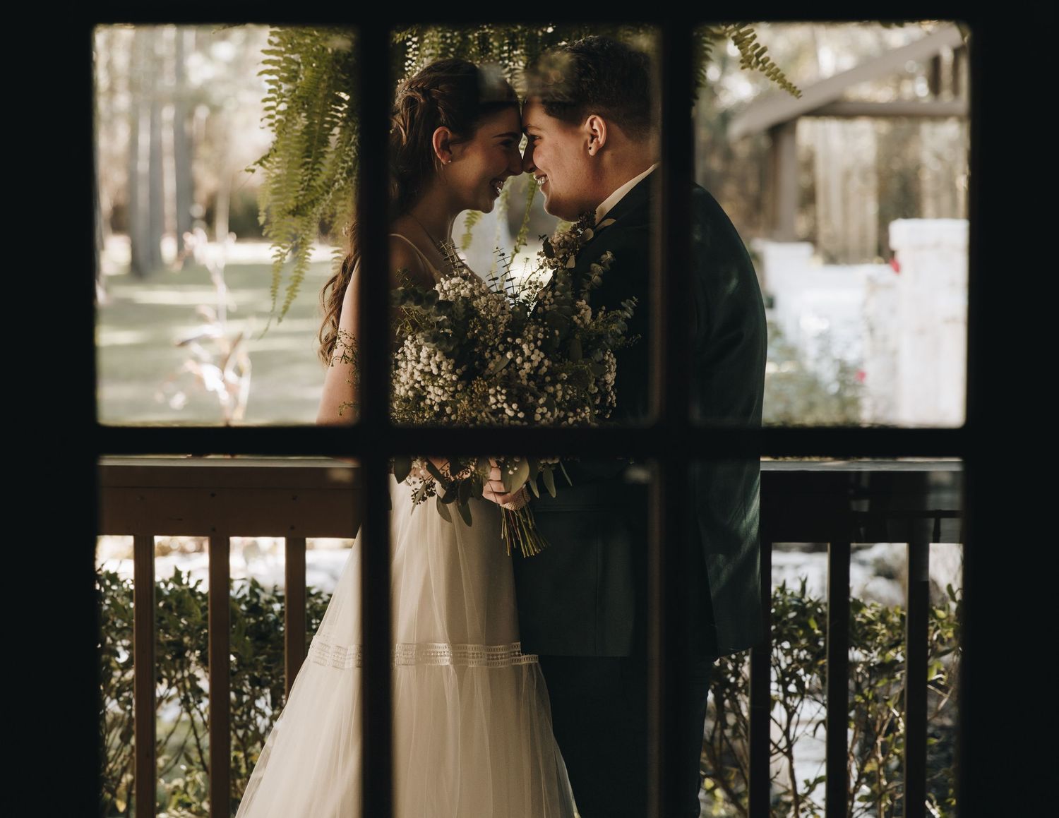 Romantic bride and groom touch foreheads while holding a lush bouquet, framed beautifully through a window pane.