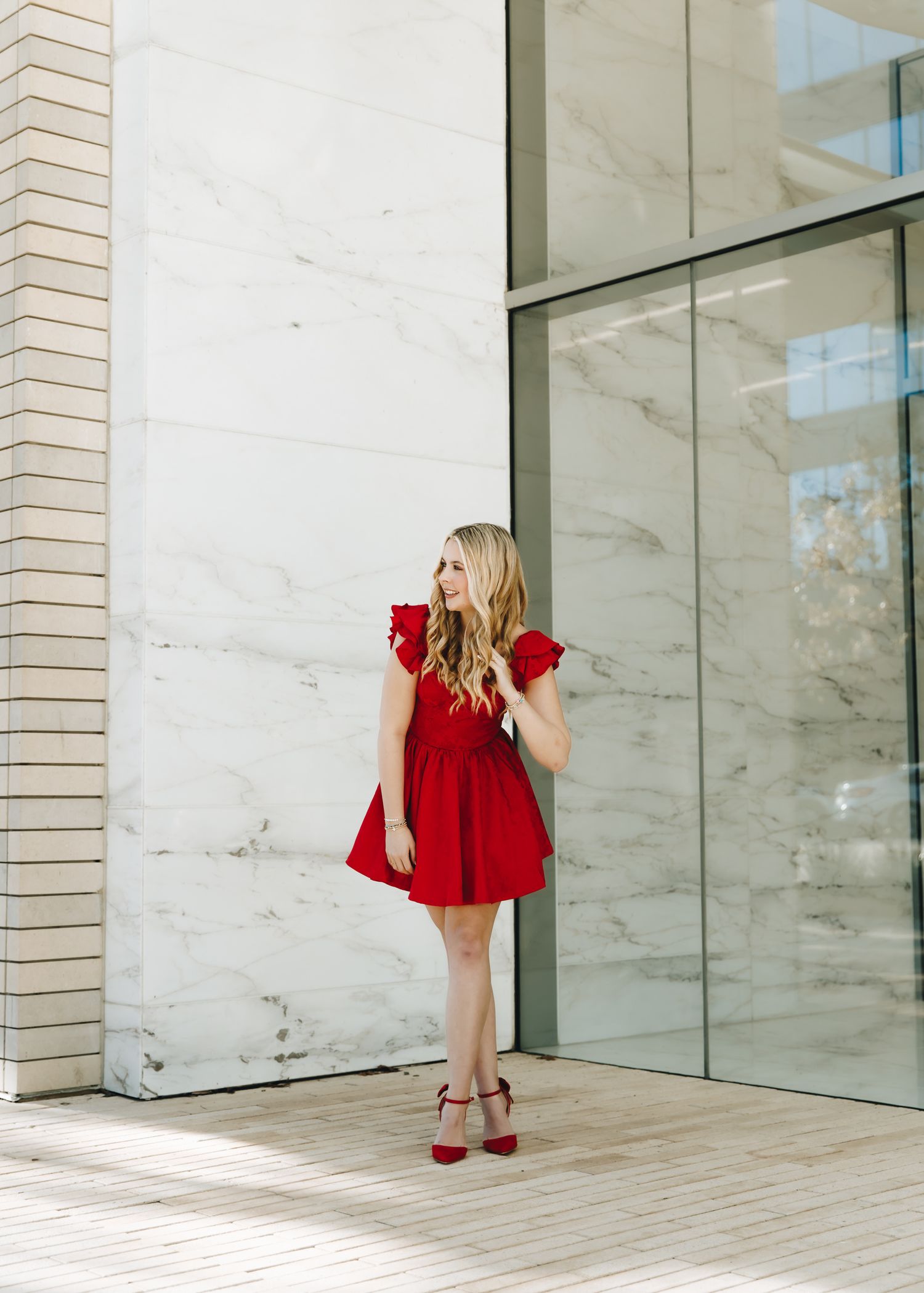 Blonde woman in red ruffled mini dress and red heels posing by marble wall and glass door.