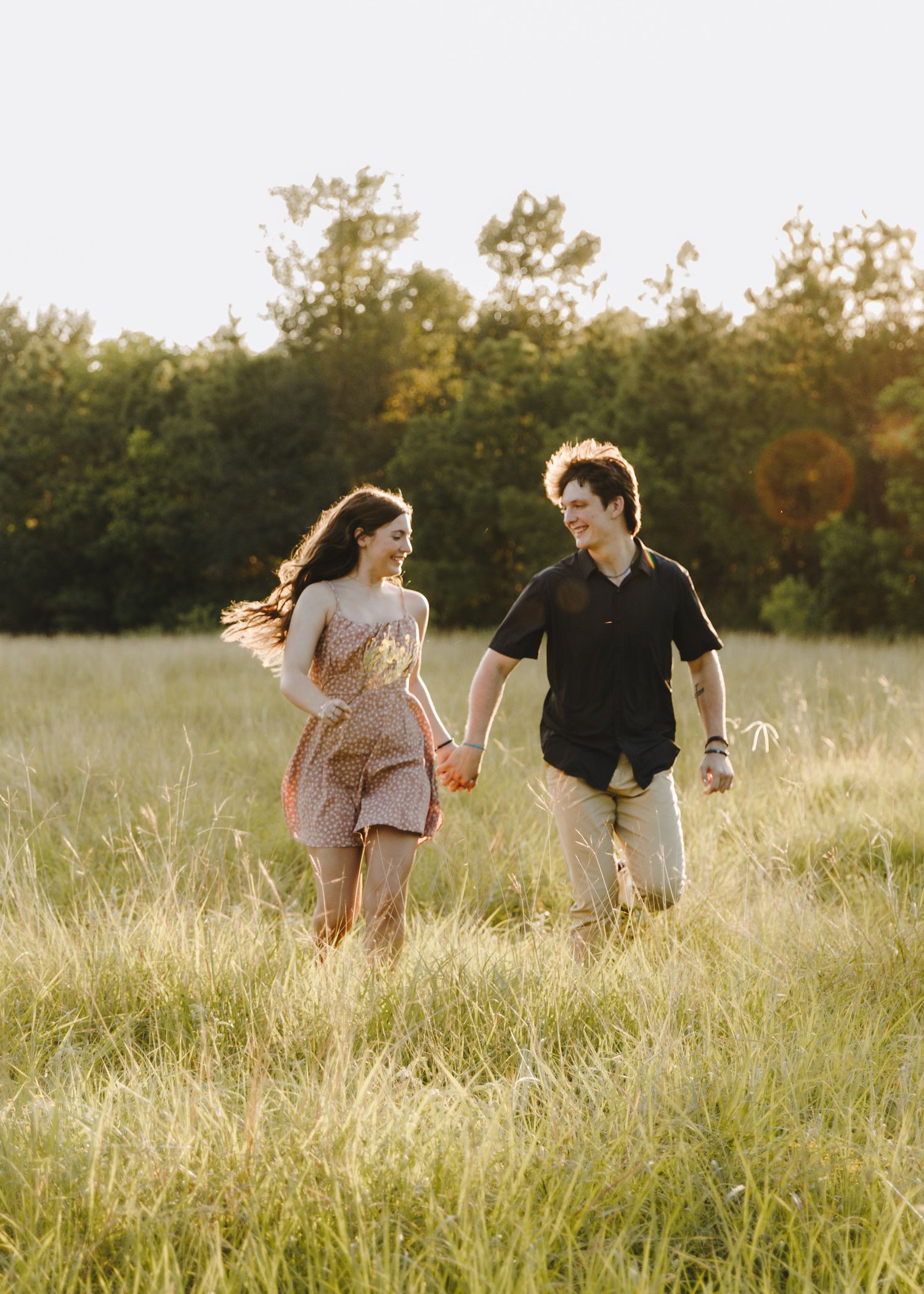 Happy couple holding hands running through a sunlit green meadow with trees in background.