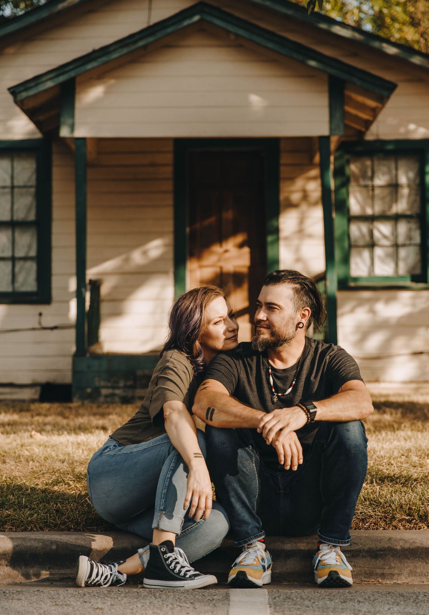 Couple sitting together on a curb, smiling at each other in front of a cottage.