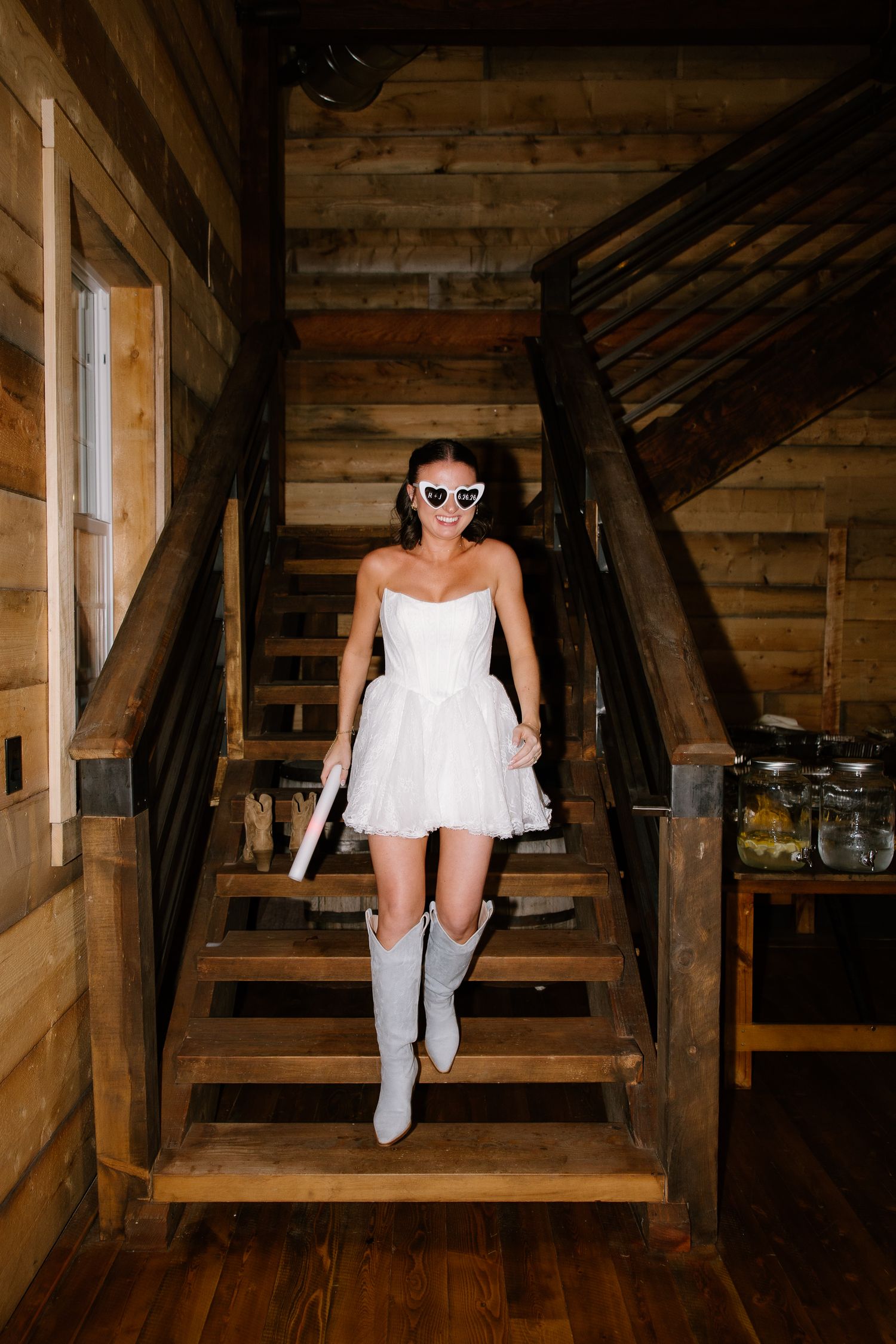 Bride in short white tulle dress, heart sunglasses, and gray cowboy boots walking down rustic wooden staircase.