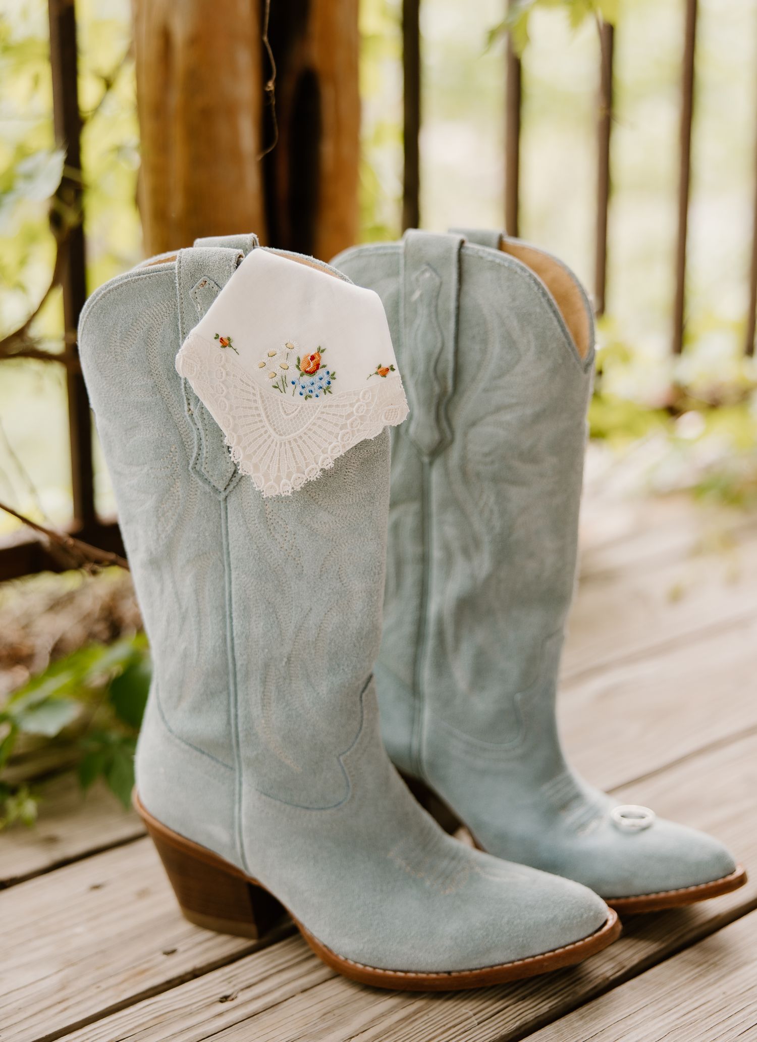 Light blue suede cowboy boots with embroidered floral handkerchief tucked inside, displayed on rustic wooden deck.