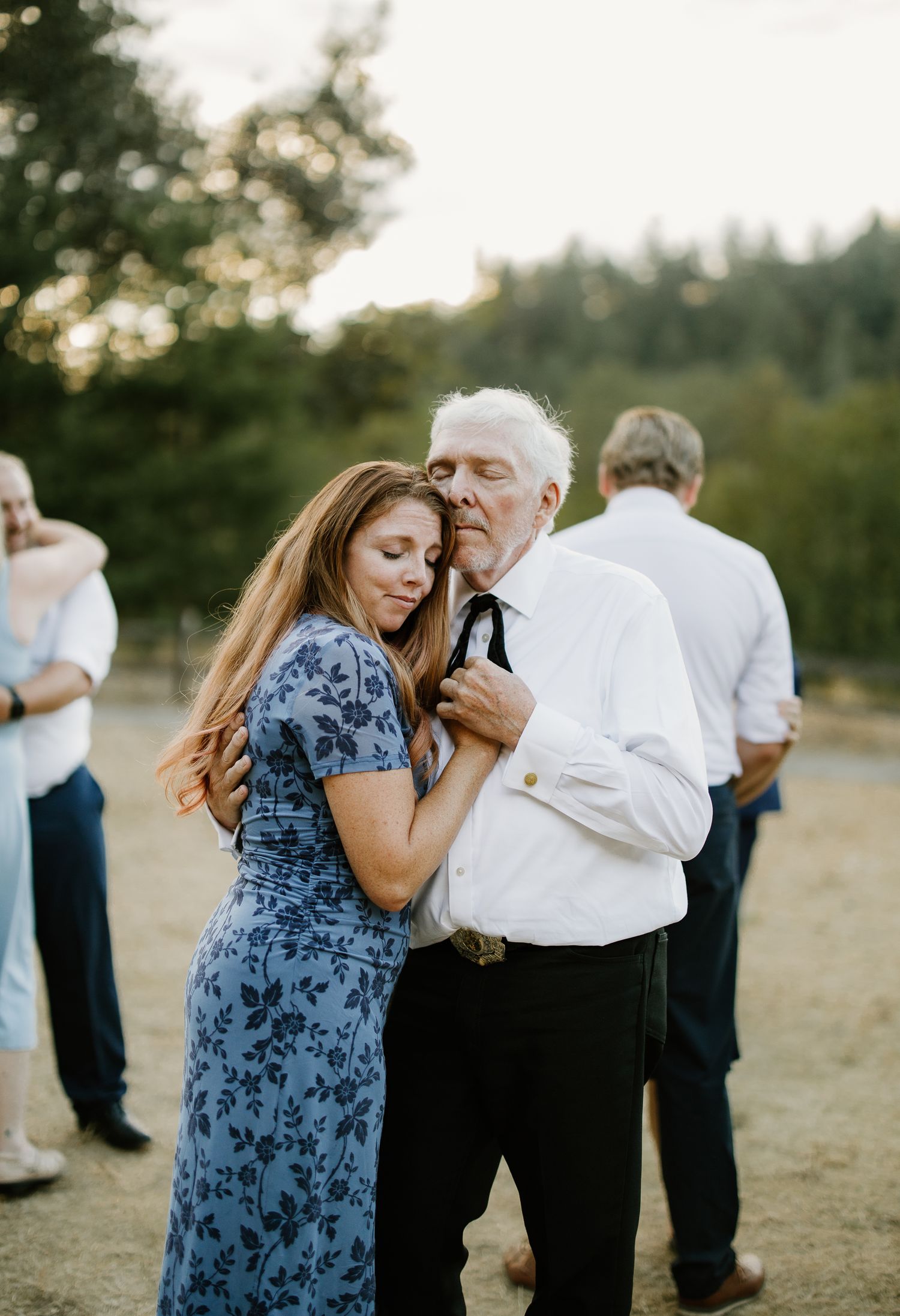 Elderly man and young woman in blue floral dress share tender dance outdoors at wedding reception.