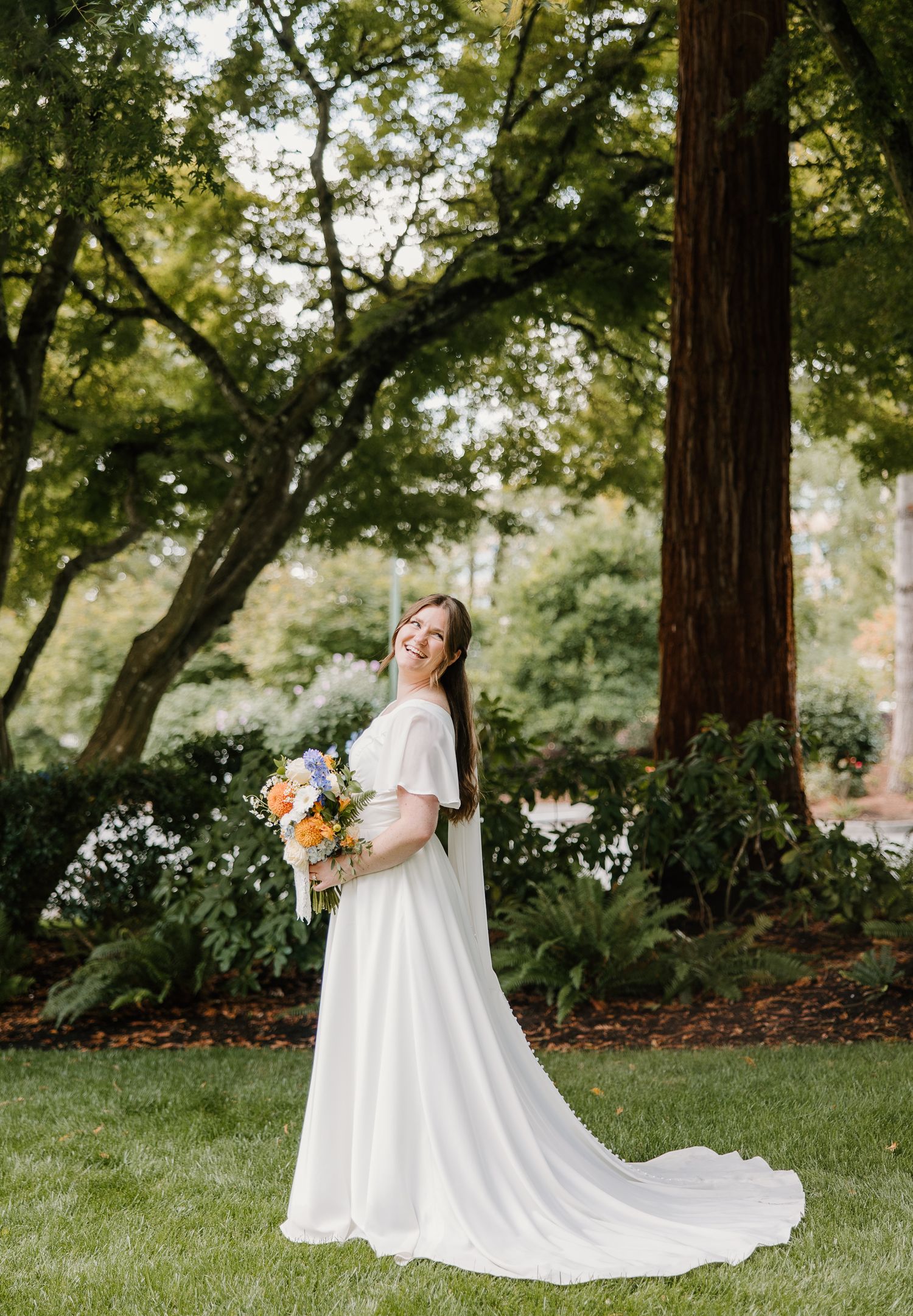 Smiling bride in white off-shoulder gown with floral bouquet stands on green lawn beside tall redwood trees.