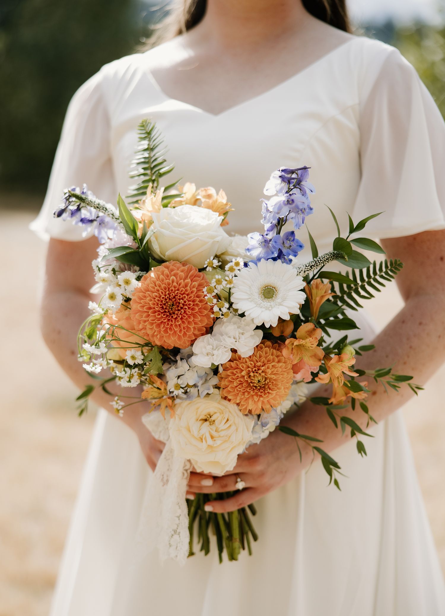 Bride in white dress holds colorful bouquet of orange dahlias, white daisies, and blue flowers.