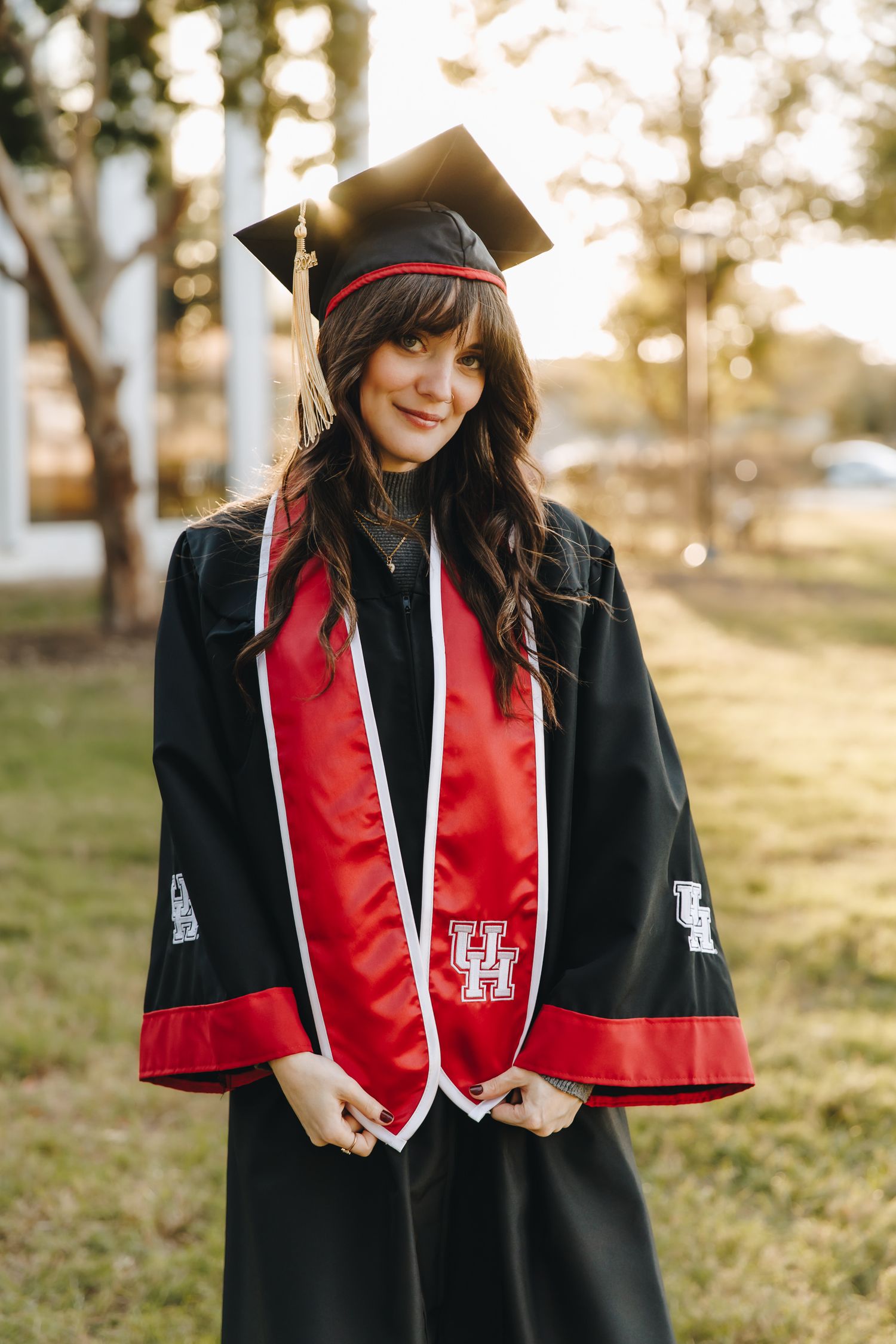 Smiling female graduate in black cap and gown with red University of Houston stole outdoors.