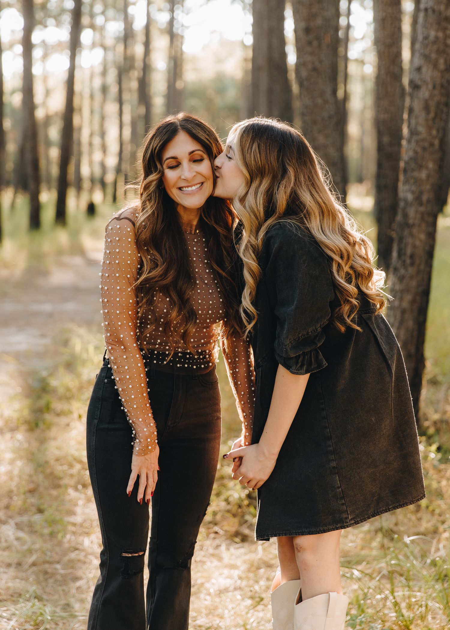 Two women with long wavy hair sharing a sweet kiss on the cheek in a pine forest.