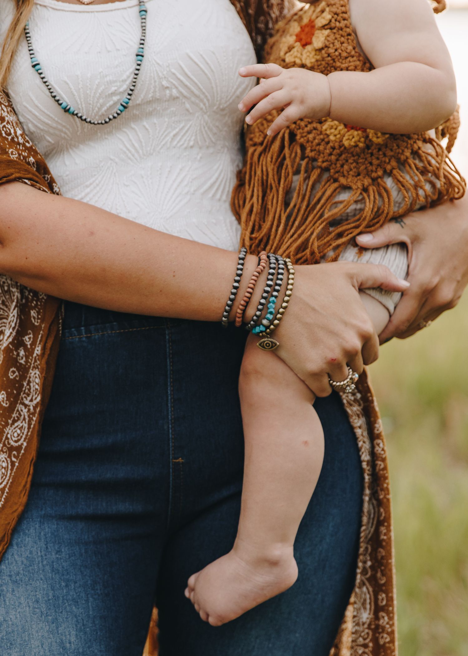 Close-up of a woman holding a baby, wearing turquoise beaded bracelets and necklace outdoors.