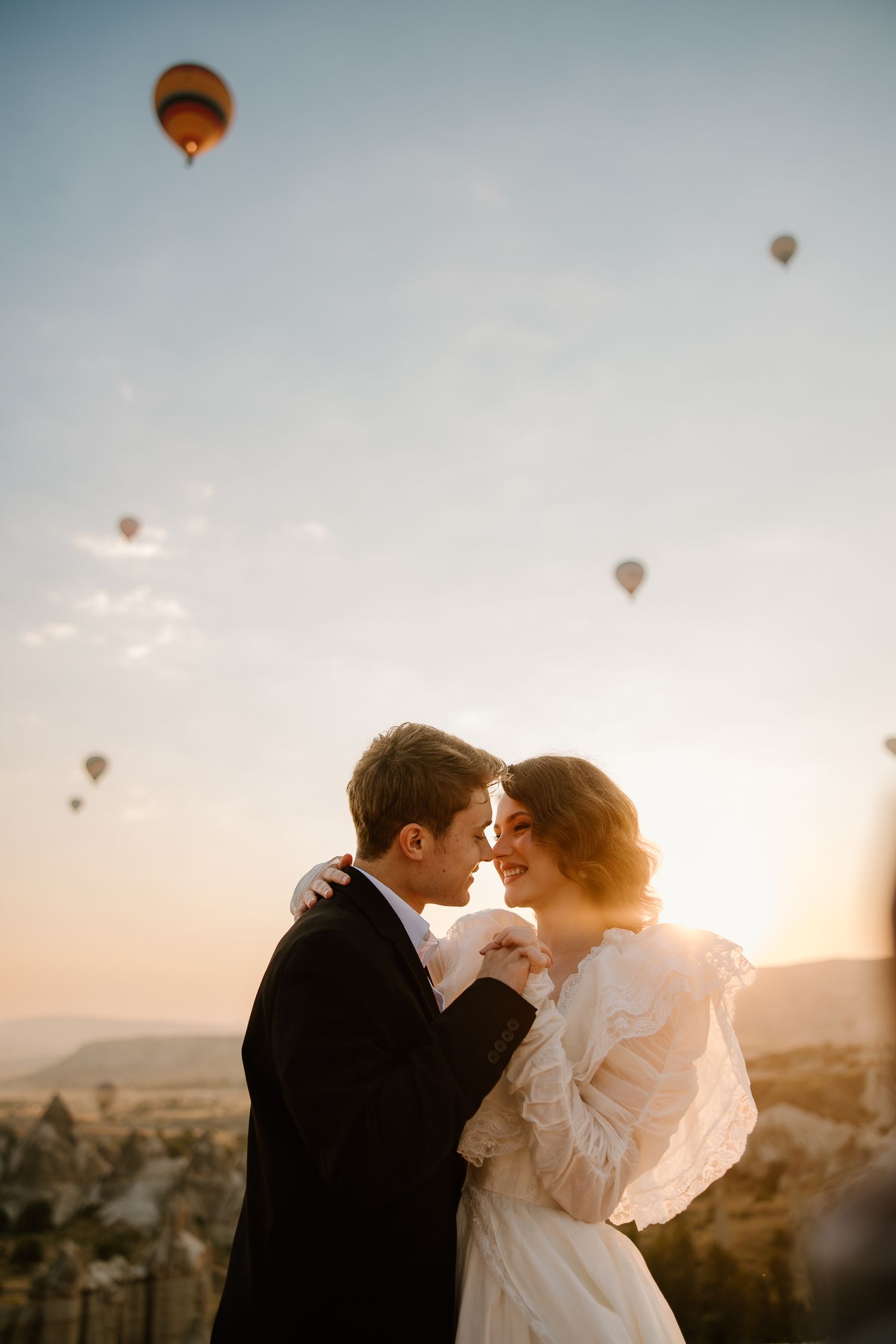 Romantic couple embracing at sunset in Cappadocia with colorful hot air balloons floating in the sky.