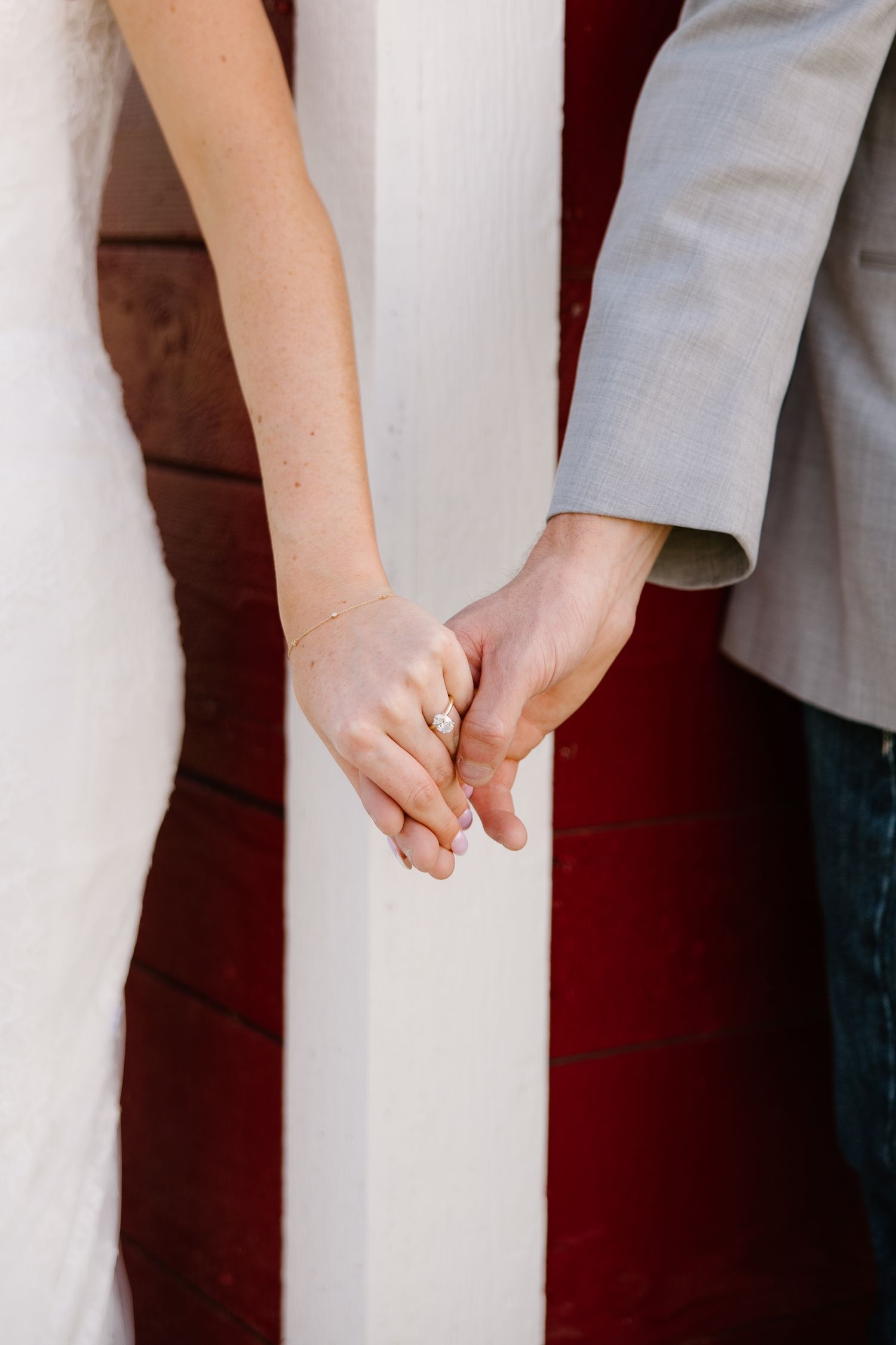 Bride and groom holding hands, showing wedding ring, standing by white pillar with red barn wall background.