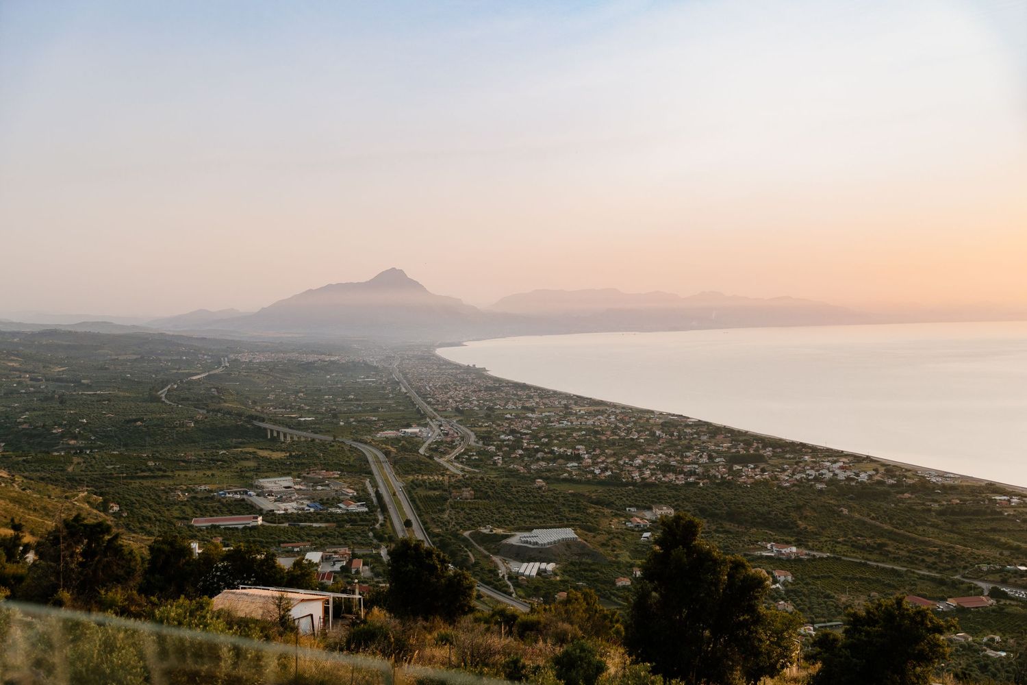 Aerial sunset view of a coastal town with a mountain backdrop and winding road along the shoreline.