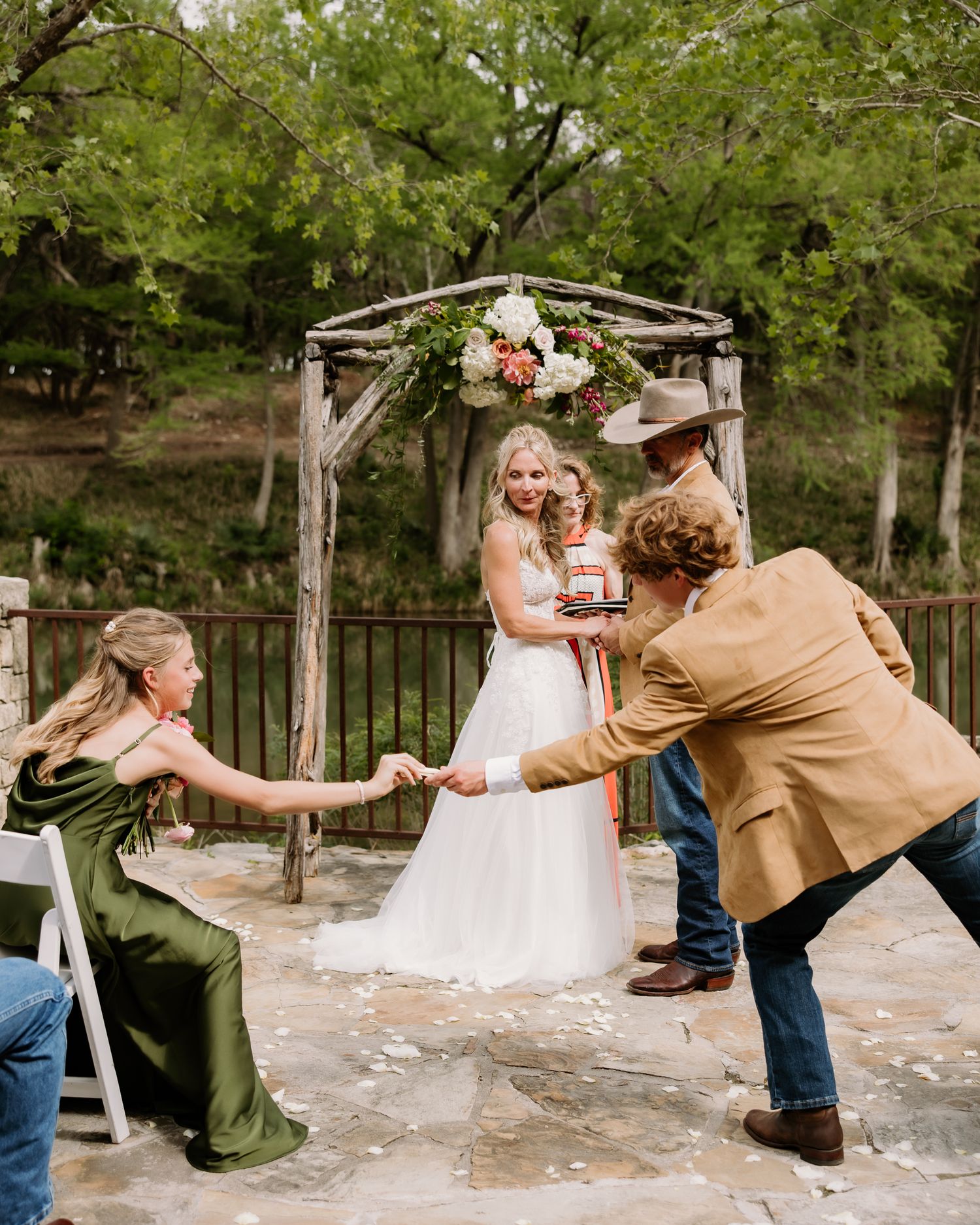 Bride in white gown and groom in tan jacket celebrate at rustic wooden arch with floral decor.