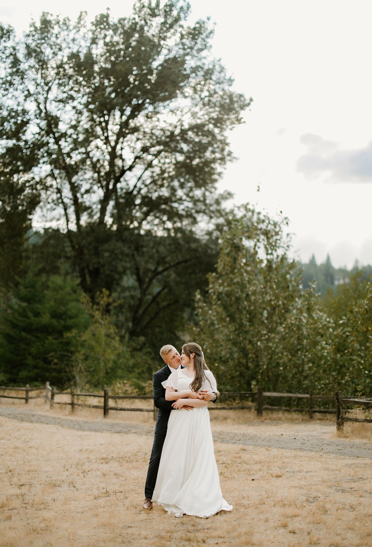 Bride and groom share a romantic kiss outdoors surrounded by trees at a rustic wedding venue.