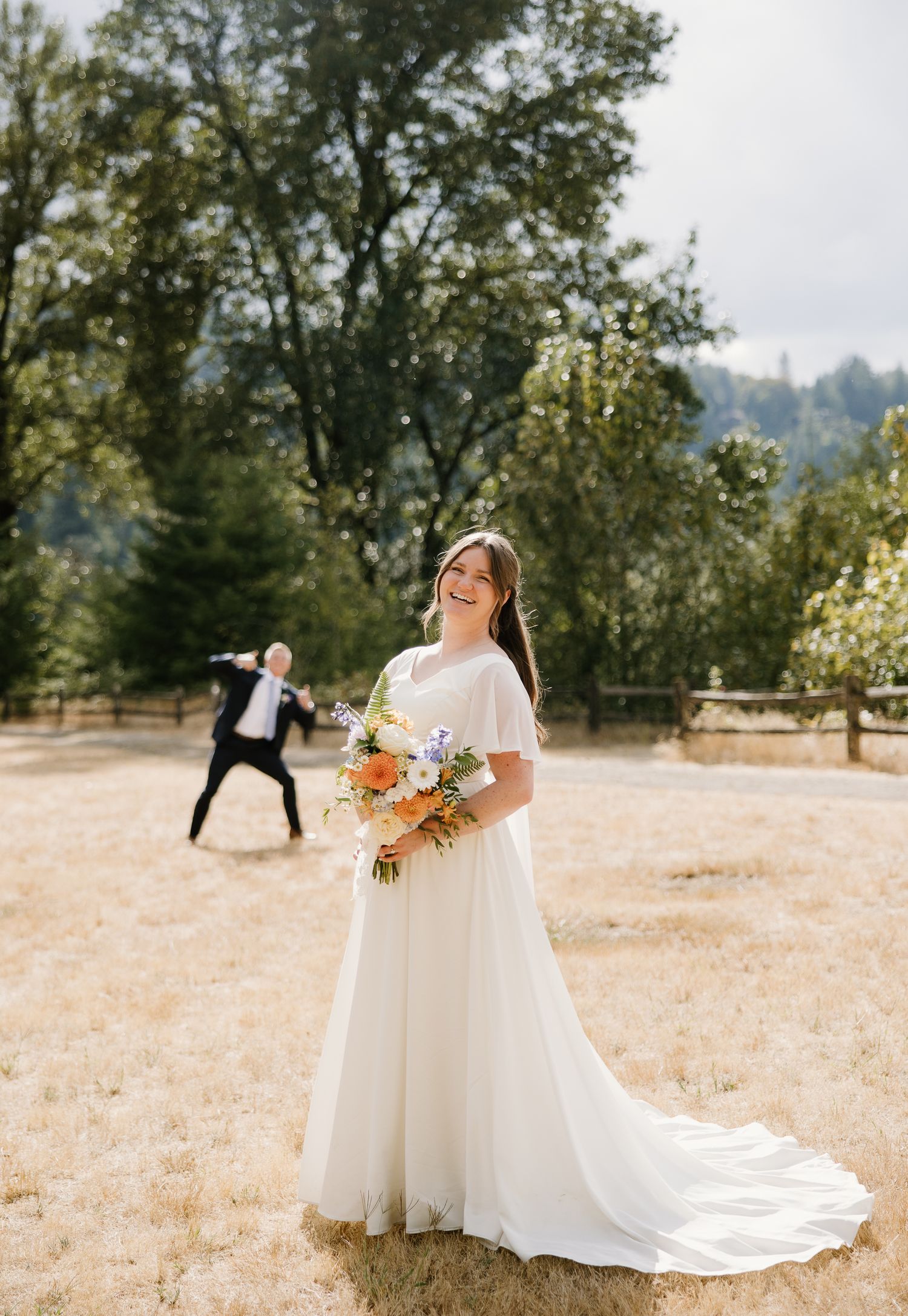 Smiling bride in white gown holds colorful bouquet while groom playfully poses in background.