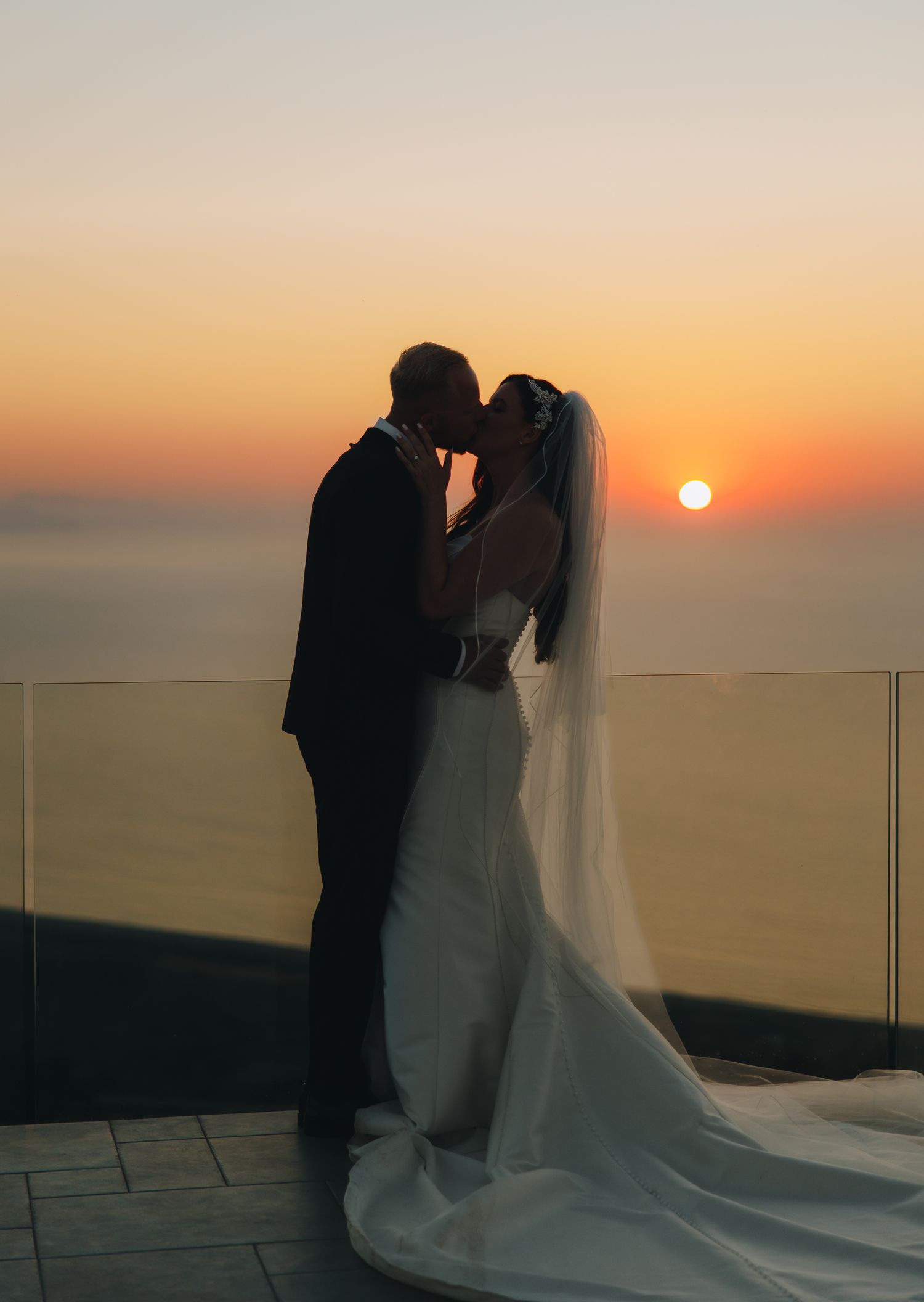 Silhouette of bride and groom sharing romantic sunset kiss with ocean horizon and golden sky backdrop.