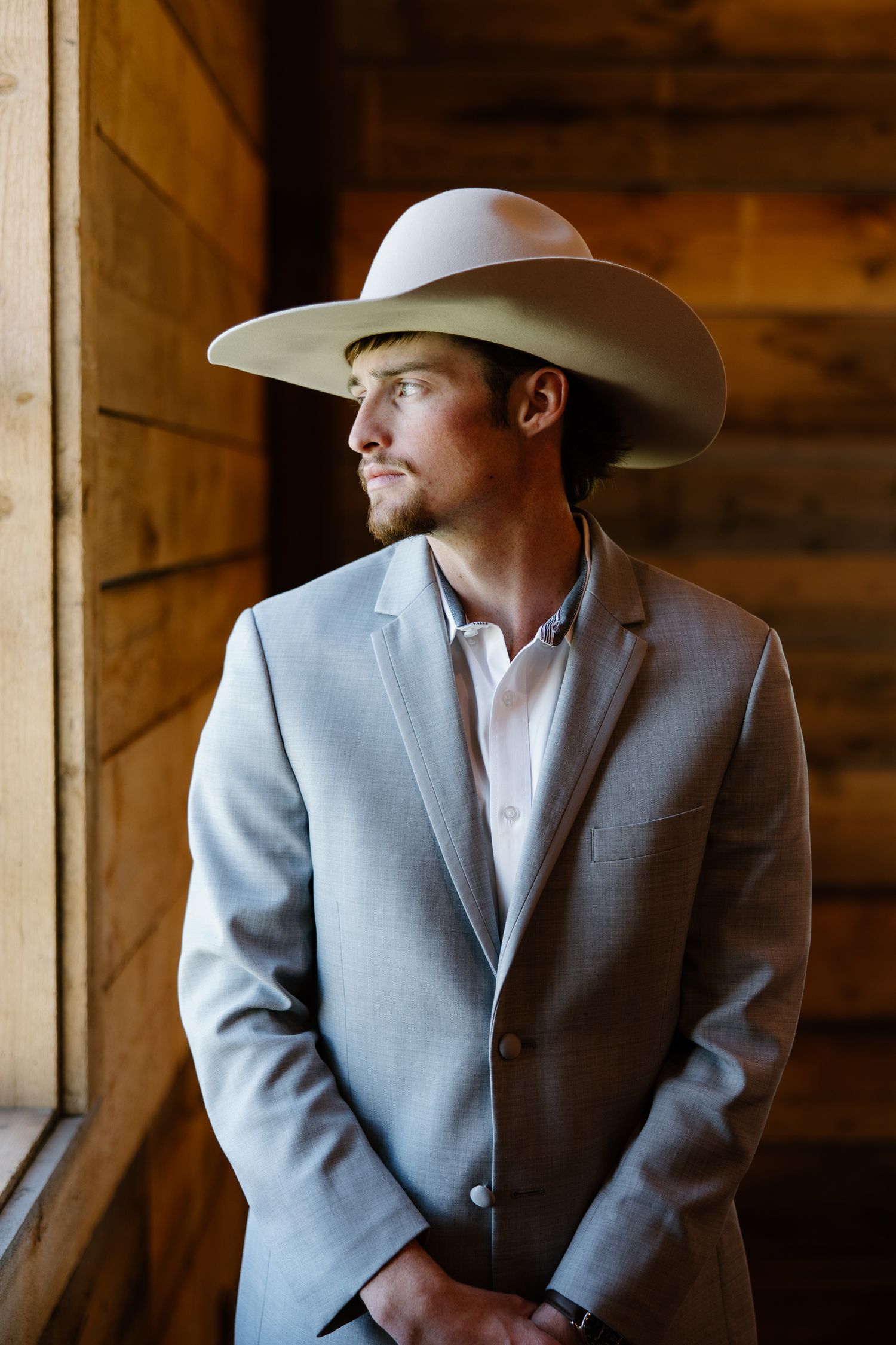 Groom in gray suit and wide-brim cowboy hat gazing out window in rustic wooden barn.