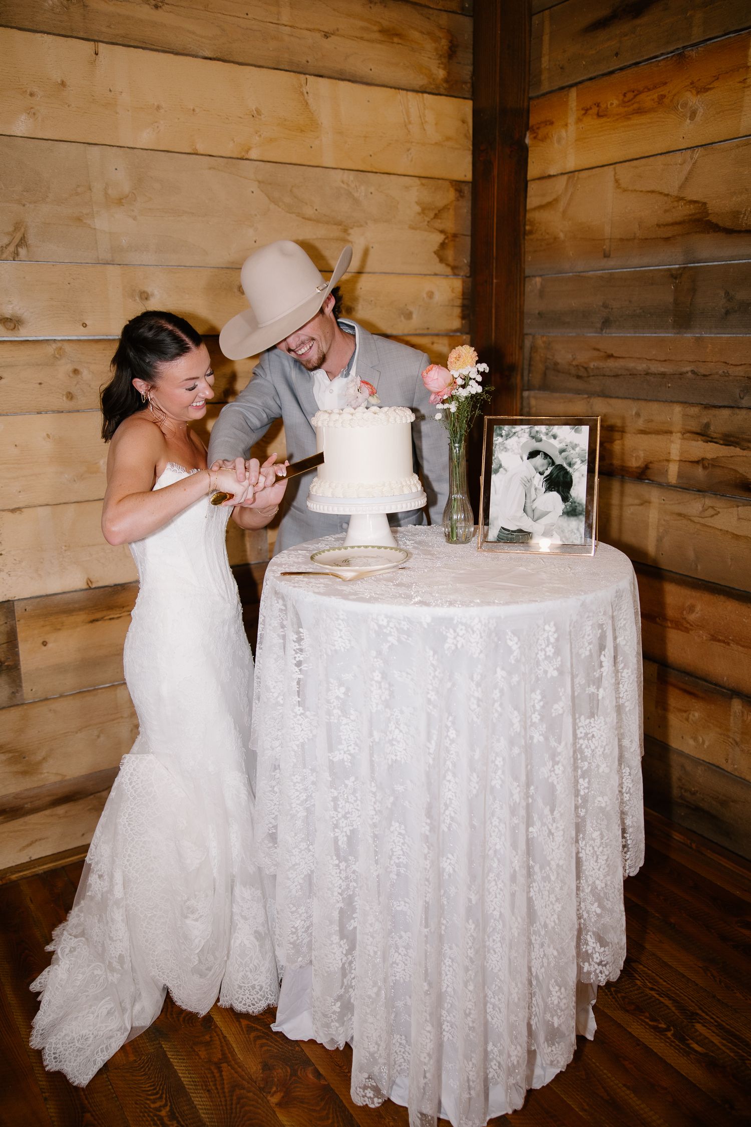 Bride and groom in cowboy hat cutting white wedding cake at rustic barn reception.