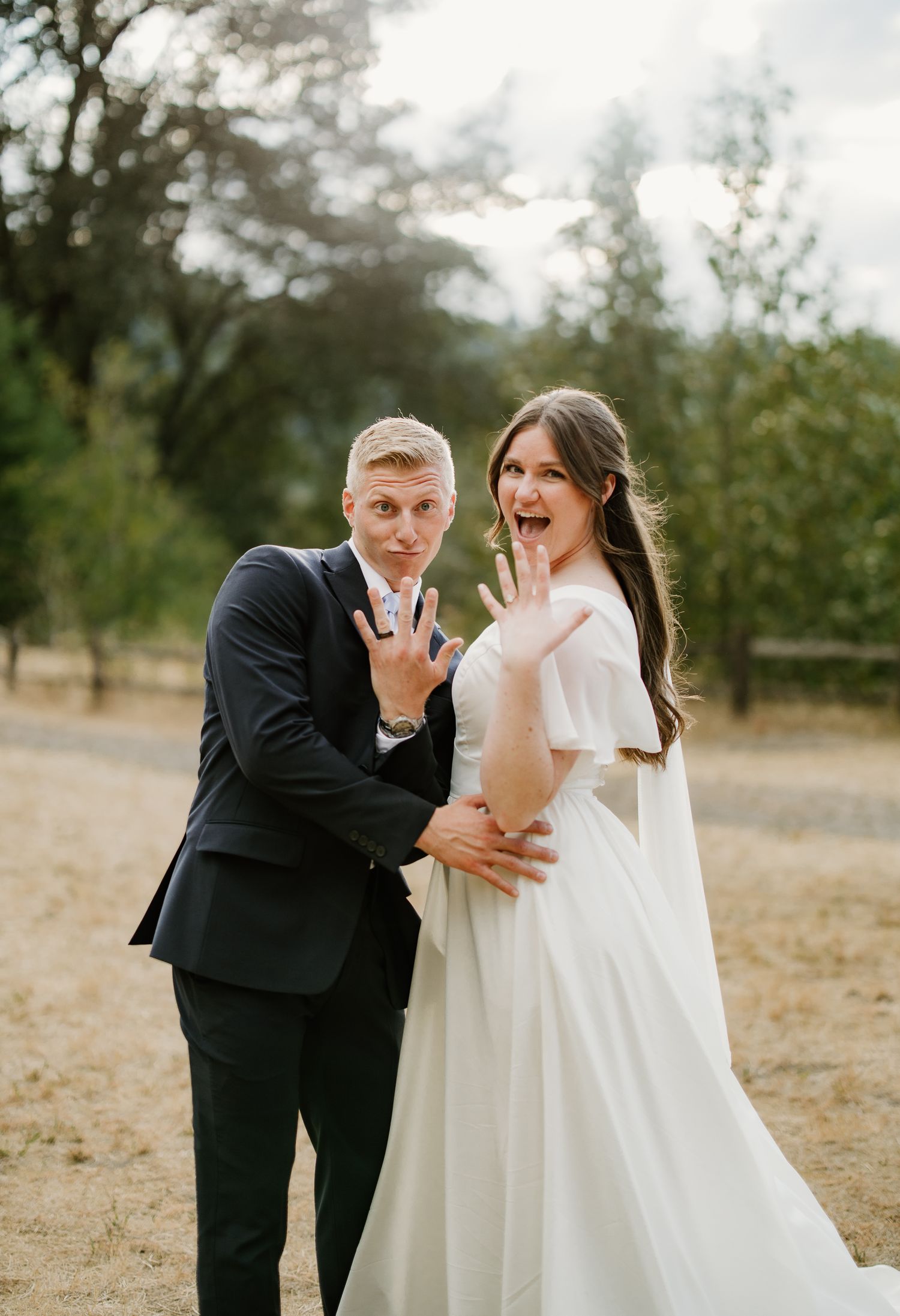 Excited bride and groom show off wedding rings while posing playfully in an outdoor rustic setting.