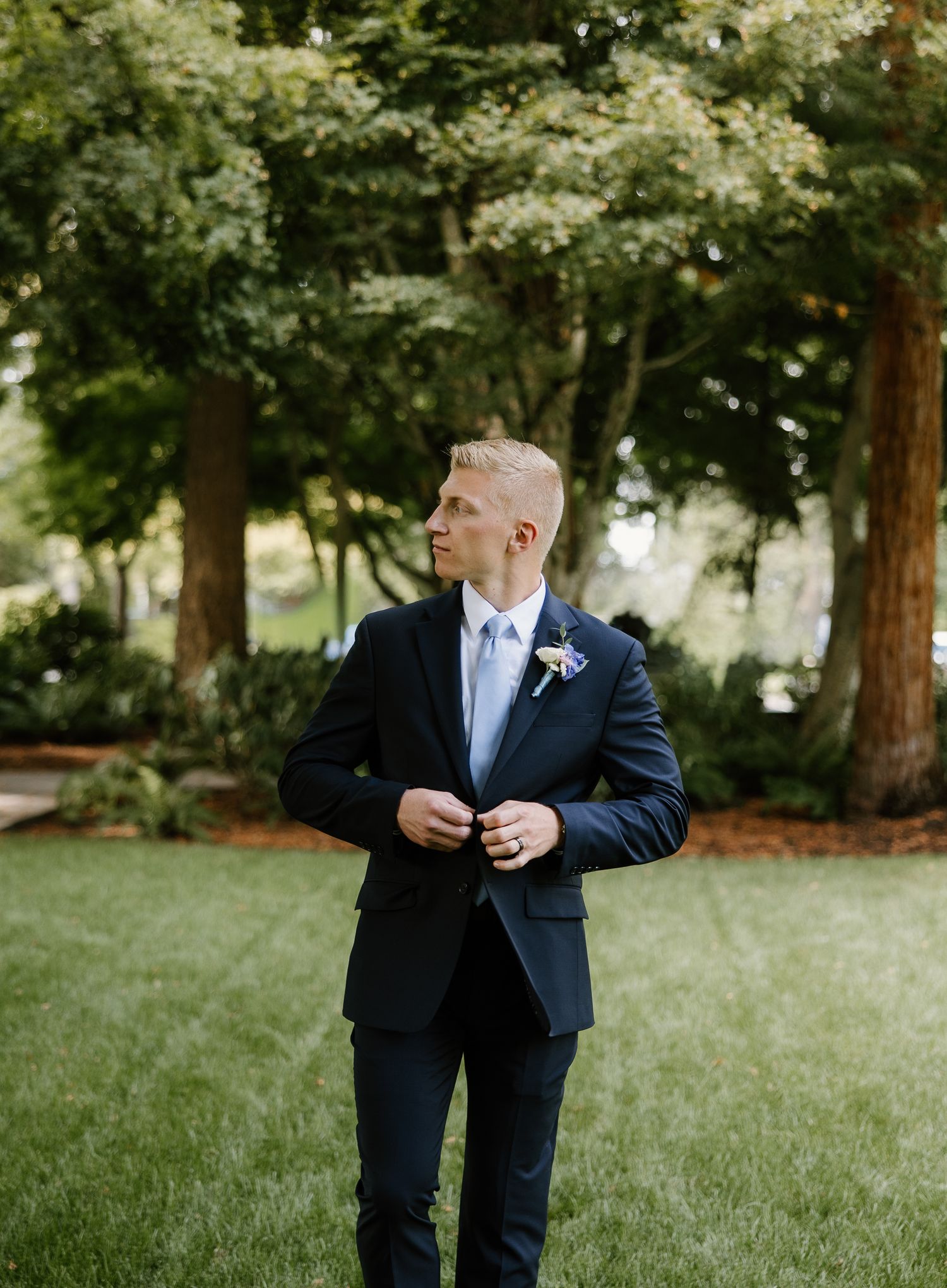 Dapper groom in navy suit with boutonniere buttons his jacket on a green lawn outdoors.