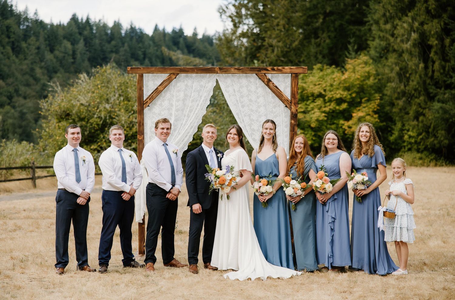 Wedding party poses before rustic wooden arch with white draping in golden field with forest backdrop.