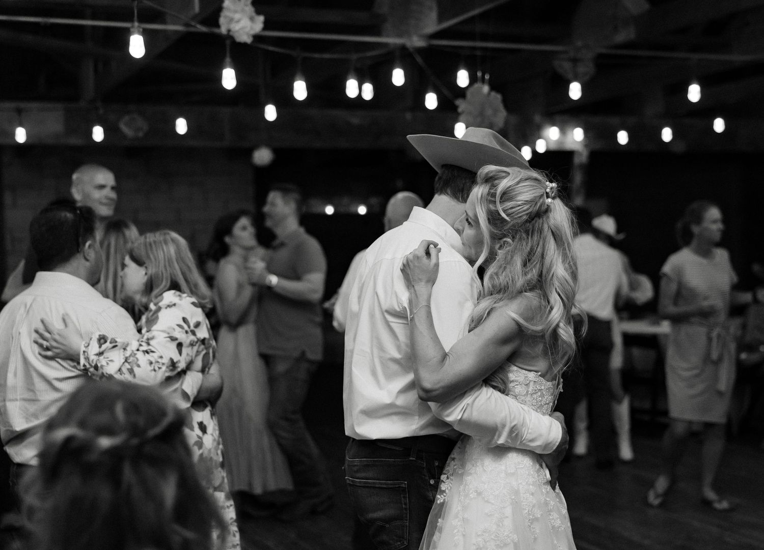 Bride and groom share a romantic first dance kiss under string lights at a rustic wedding reception venue.