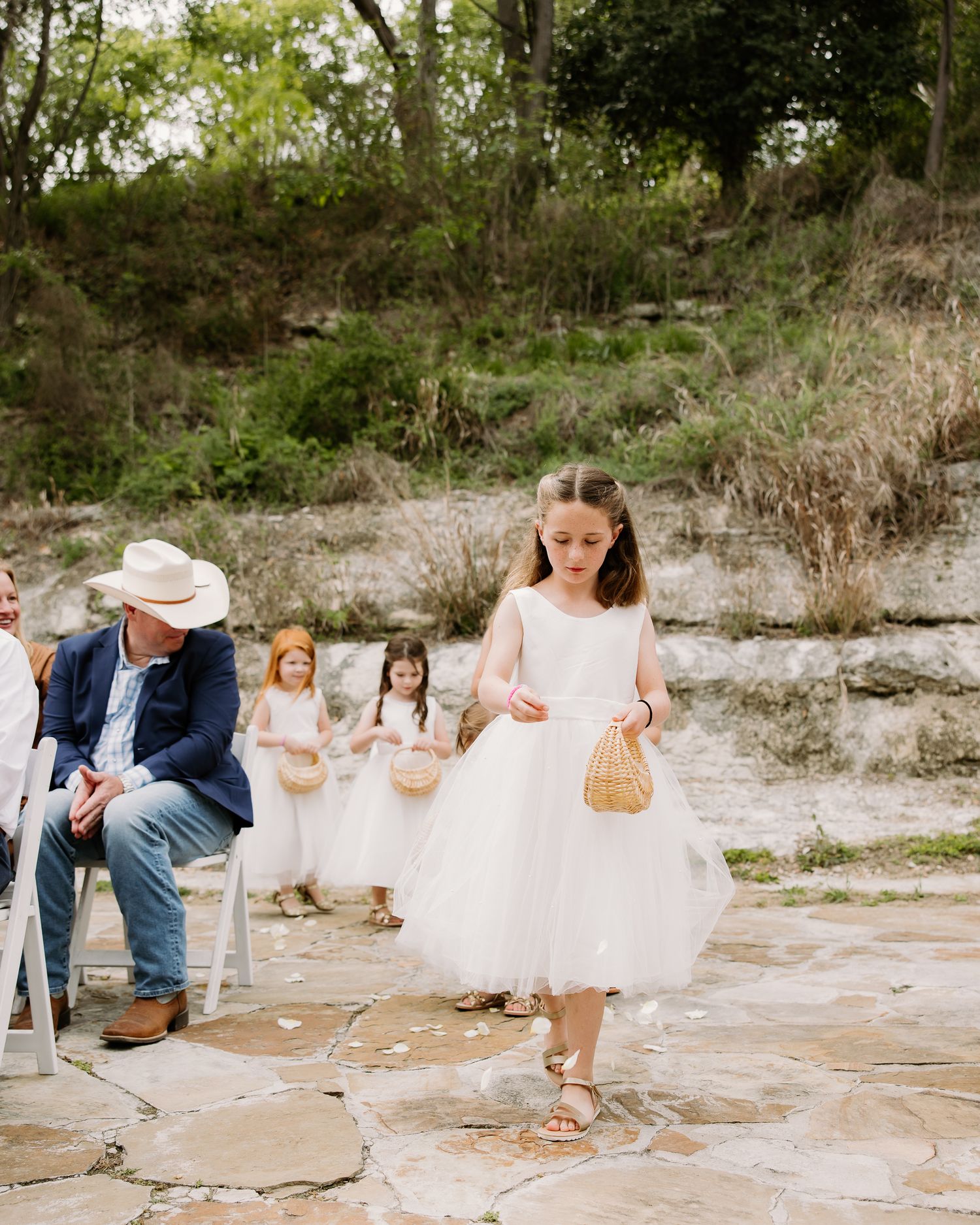 Flower girls in white tulle dresses scatter petals at an outdoor rustic wedding ceremony on a stone path.