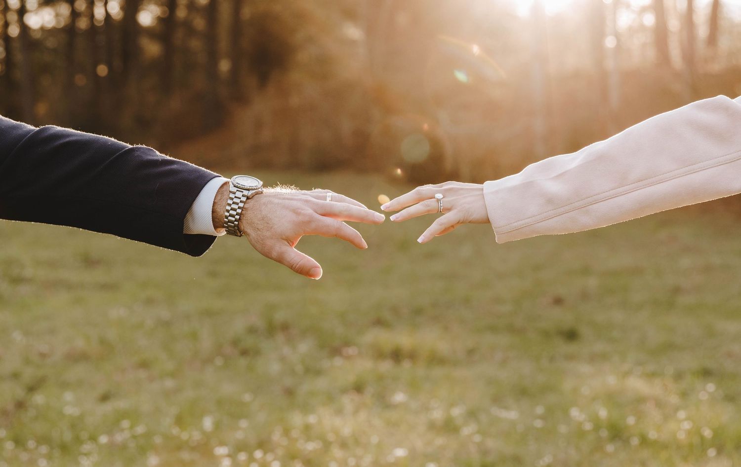 Bride and groom reach for each other's hands in a golden sunlit field during outdoor wedding photoshoot.
