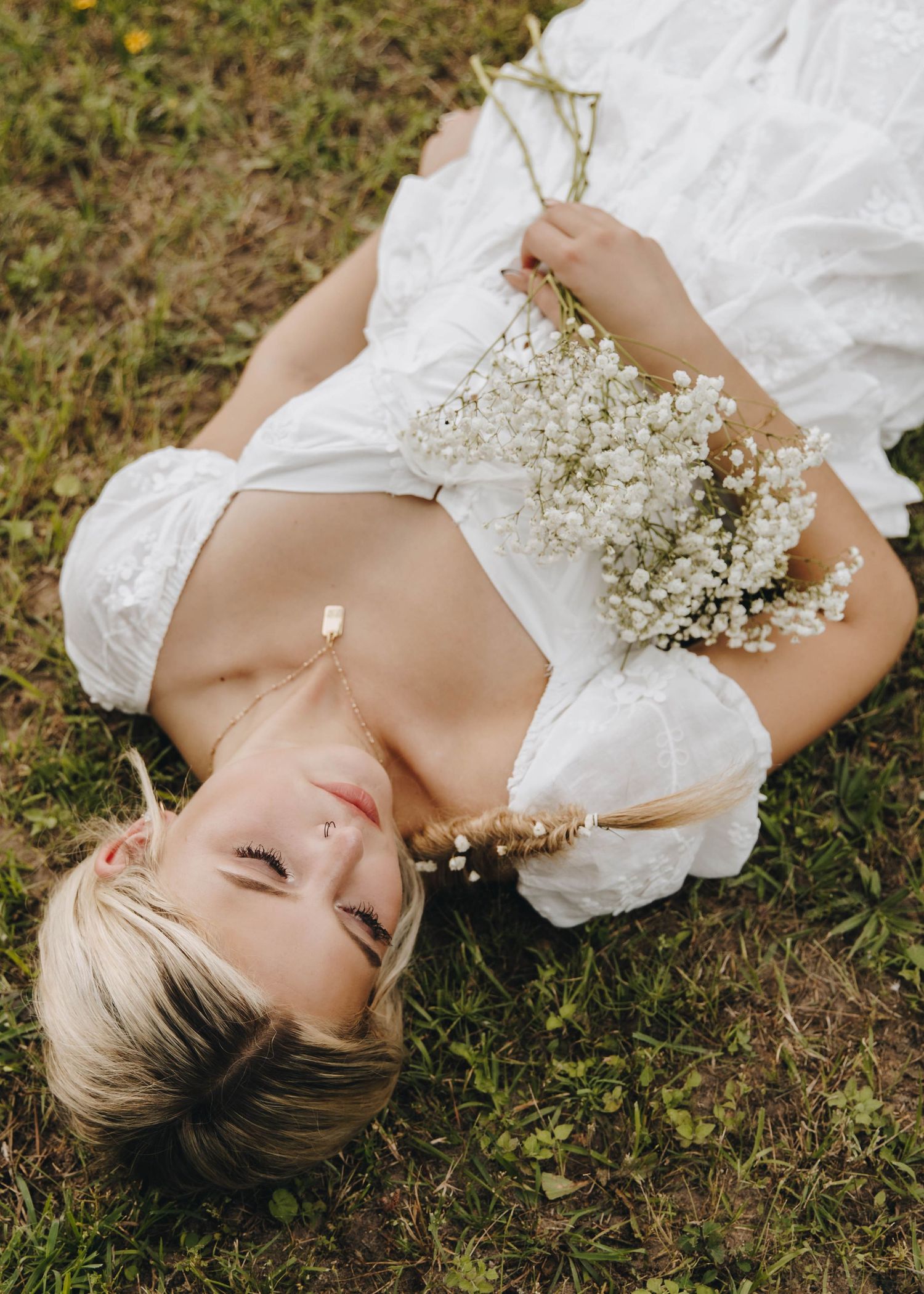 Young woman in white dress lying on grass holding delicate baby's breath flowers in a serene outdoor portrait.
