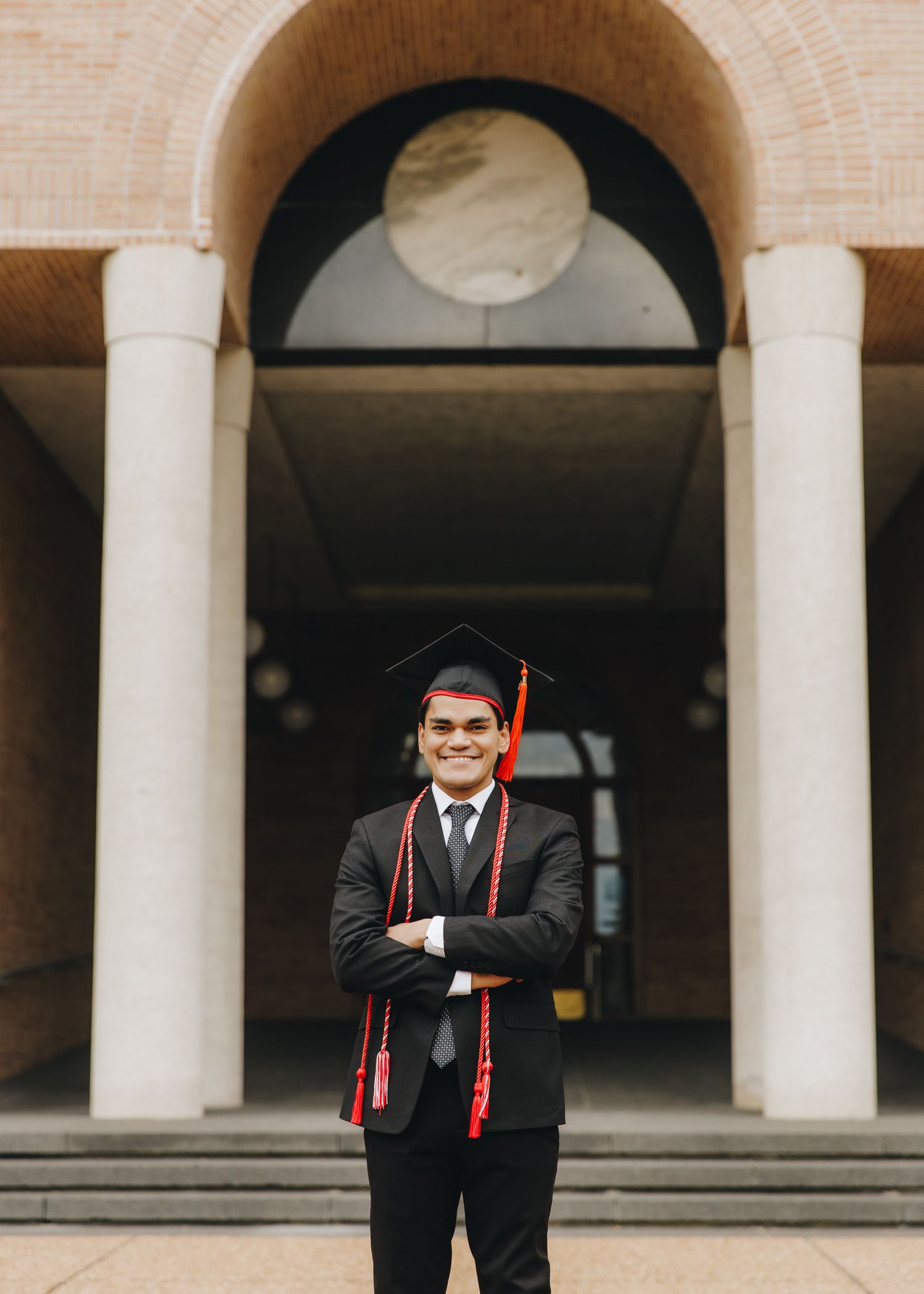 Smiling graduate in cap and gown with red stole posing arms crossed under stone archway.