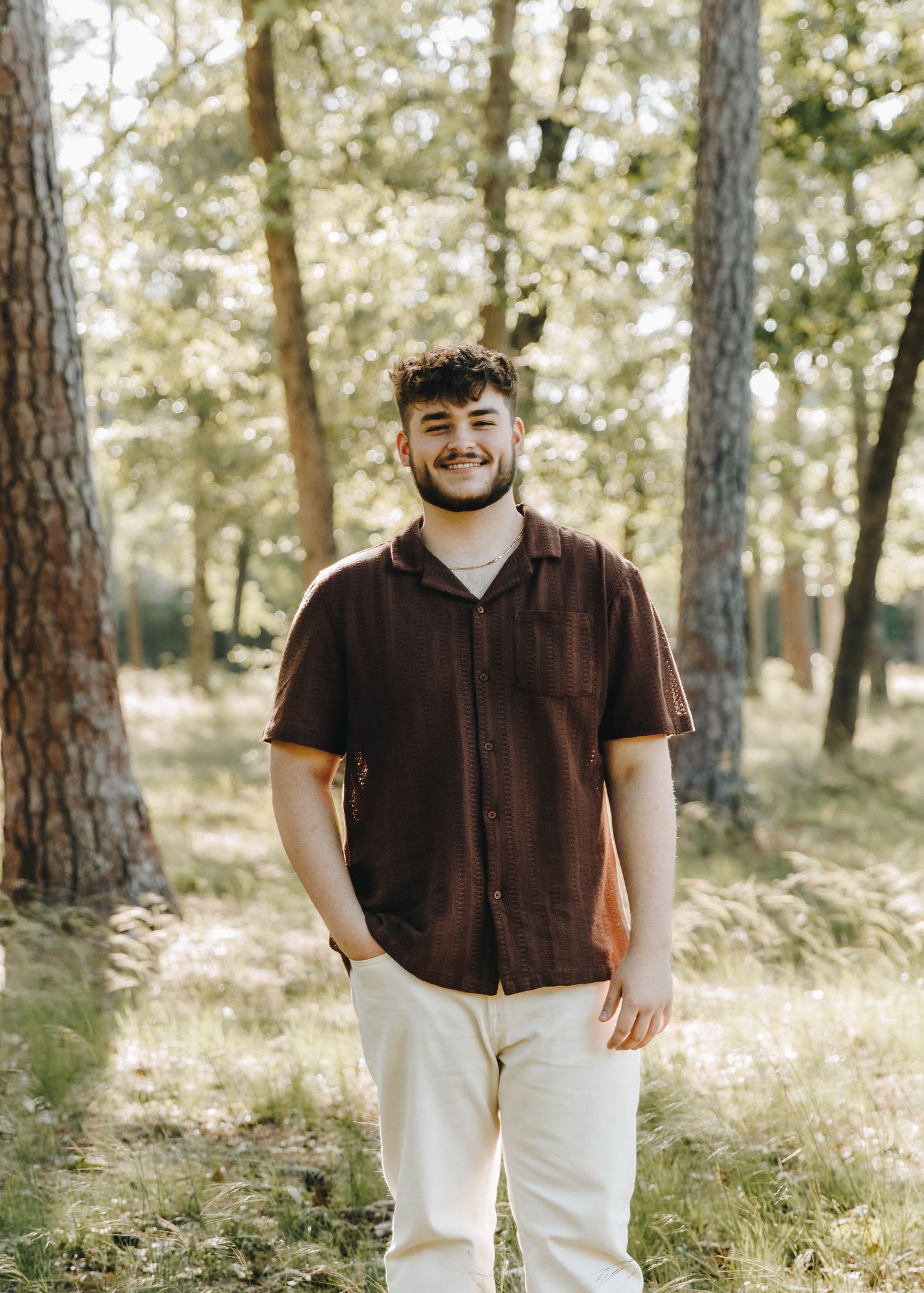 Smiling young man in brown shirt and white pants standing in a sunlit forest.