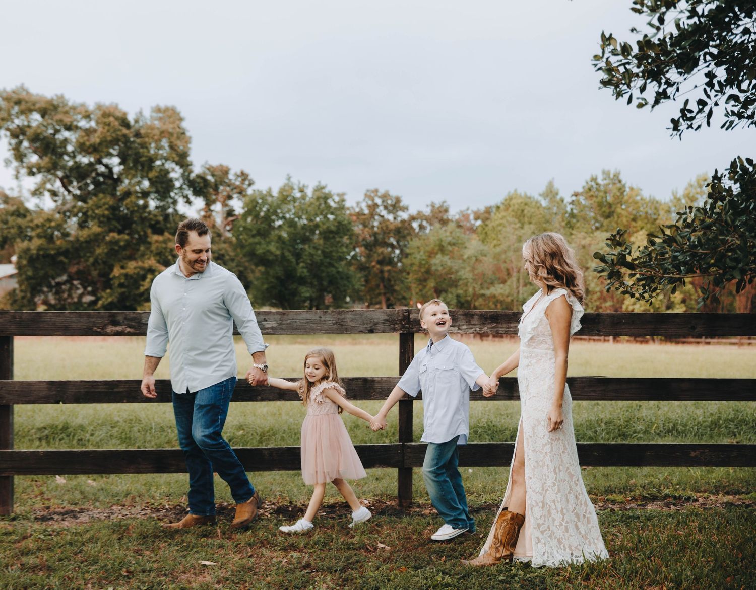 Happy family of four holds hands while walking along a wooden fence on a farm.