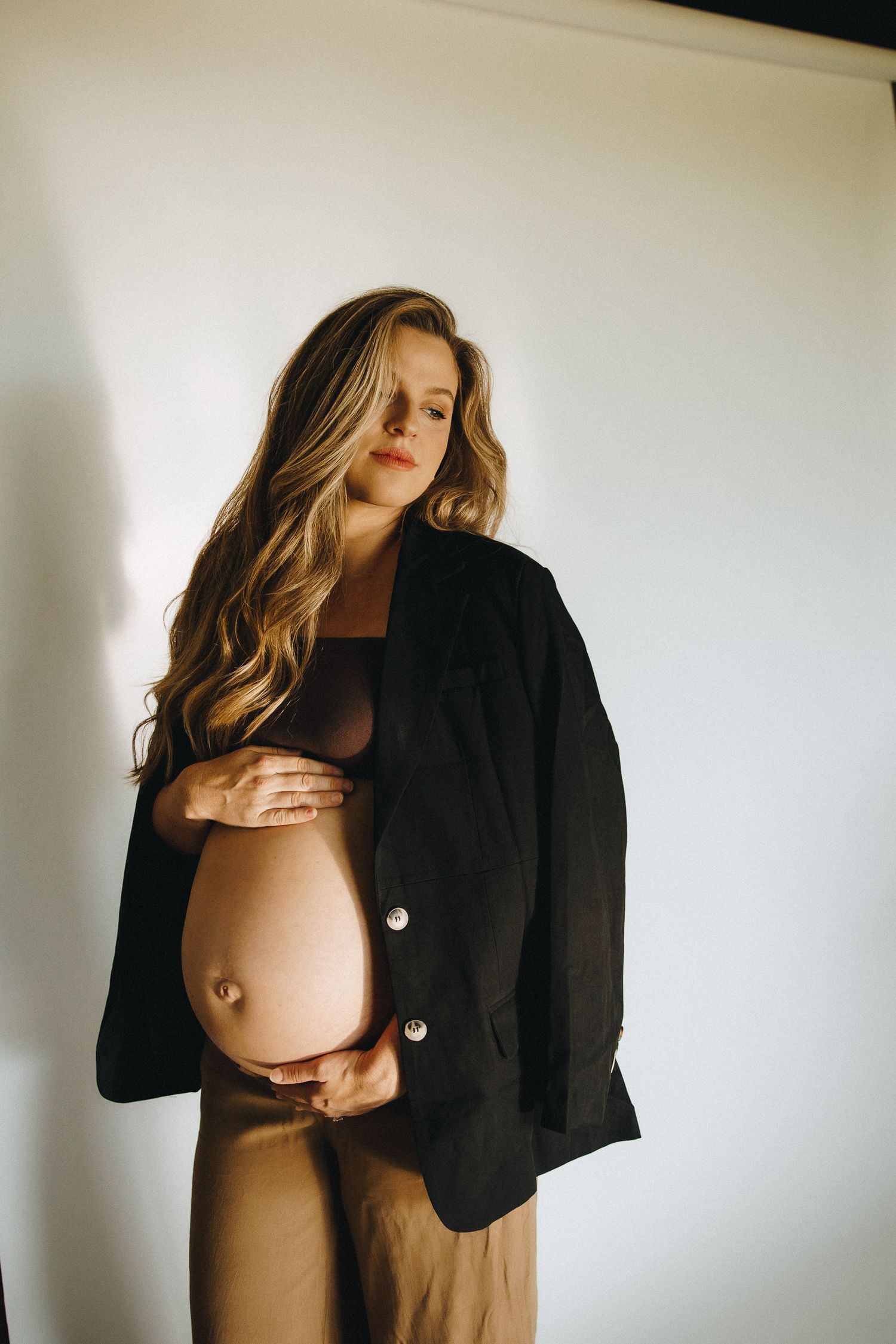 Pregnant woman in black blazer and tan outfit cradling baby bump against white background.