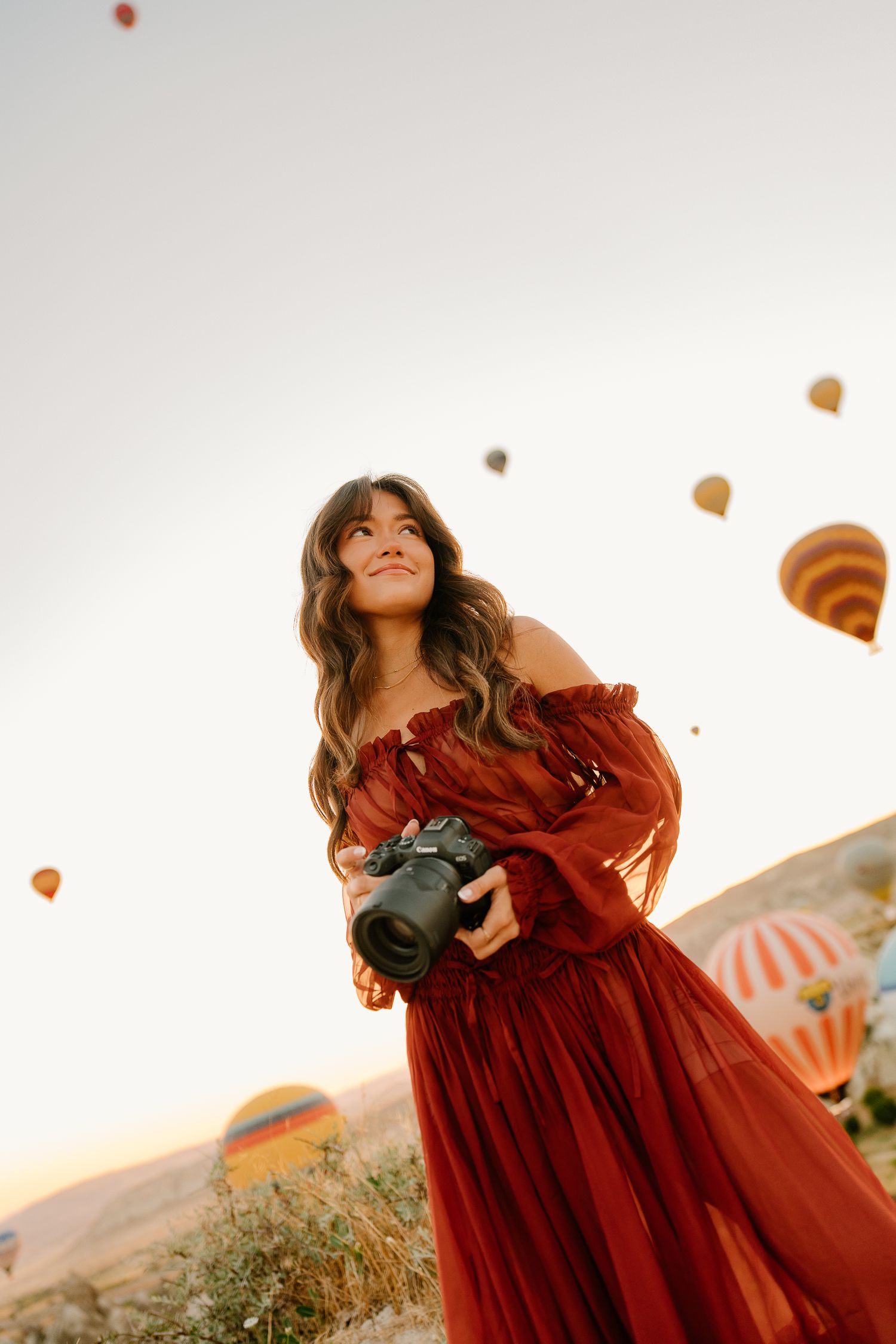Woman in red dress holding camera with hot air balloons floating in sky at sunset in Cappadocia.