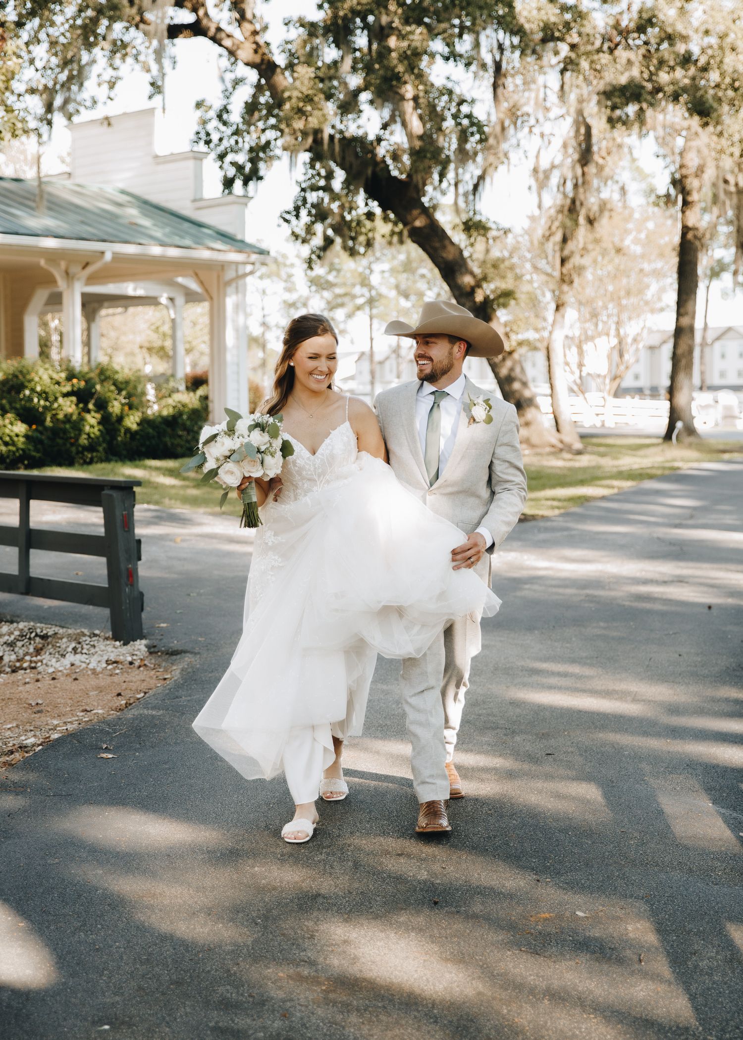 A smiling bride and groom walk together outdoors, the groom wearing a cowboy hat and tan suit.