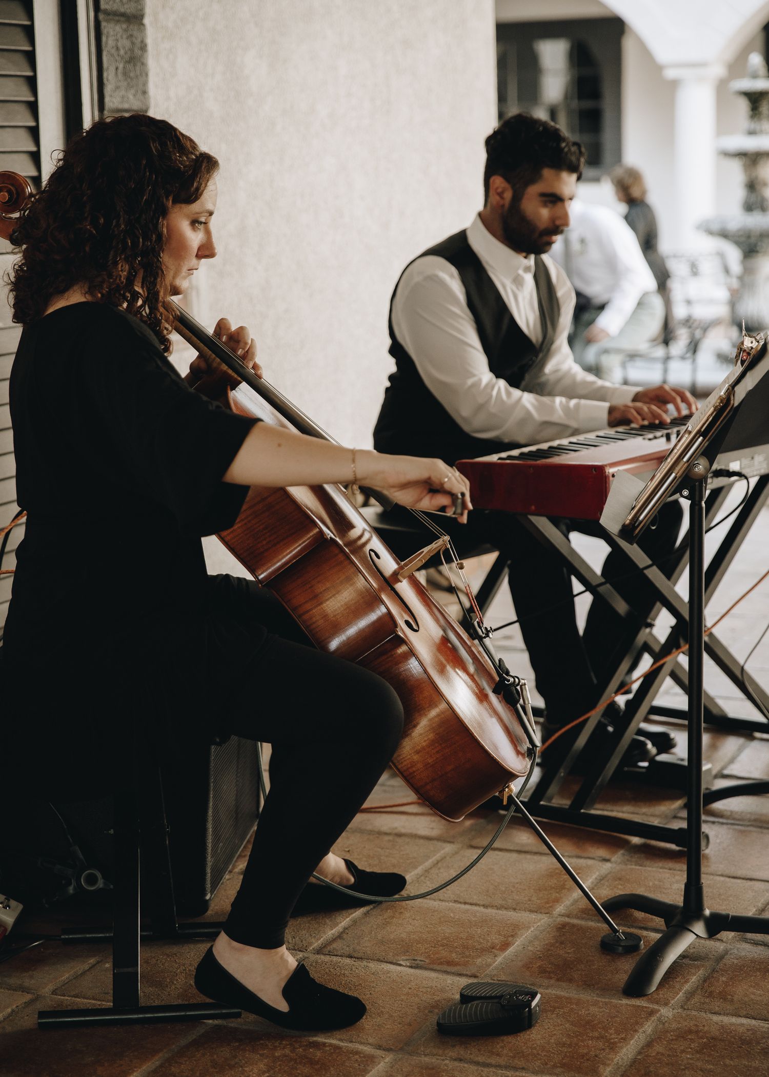 Female cellist and male keyboard player performing live music at an outdoor wedding reception under an archway.