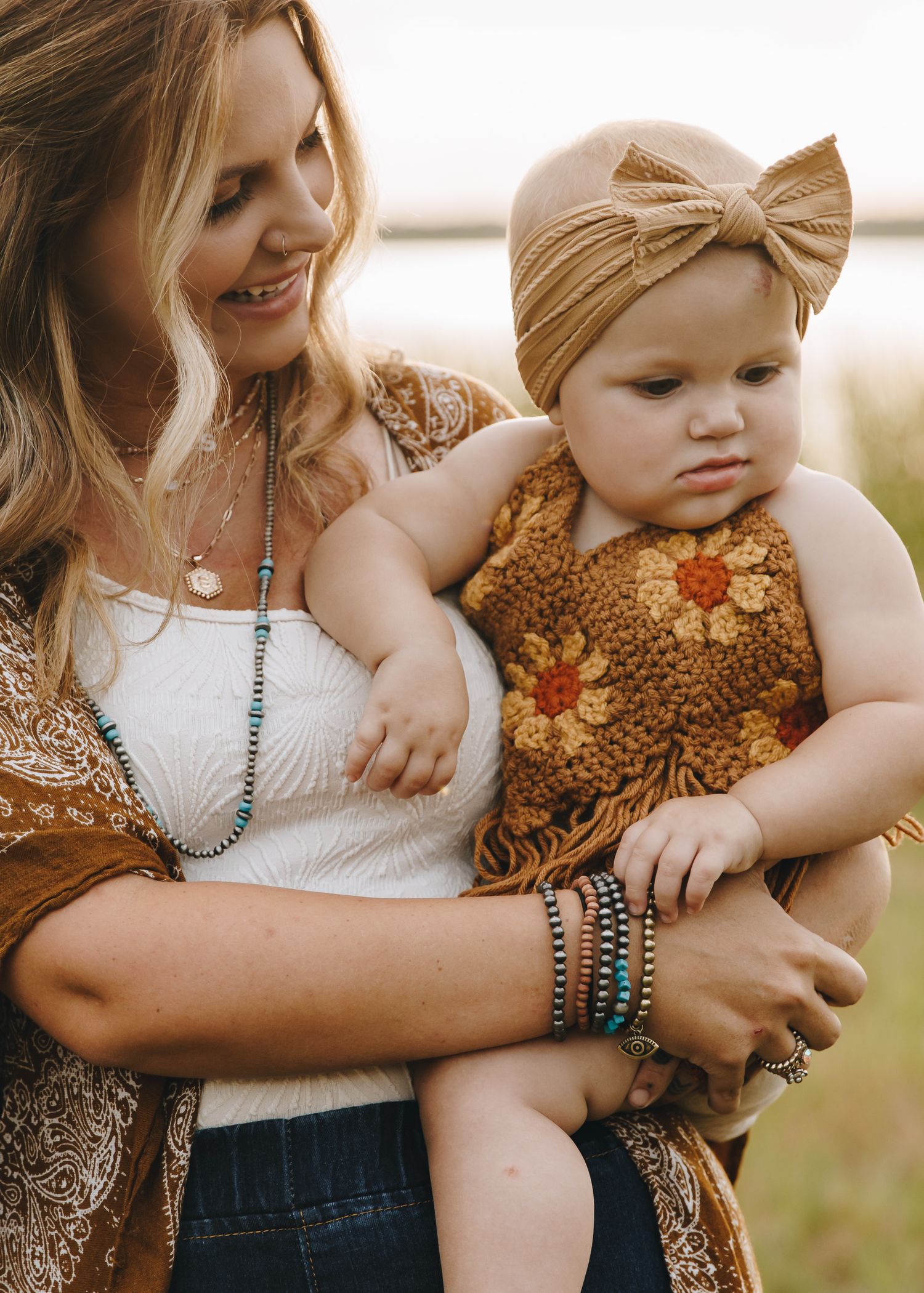 Smiling mother holding baby in mustard crochet outfit with bow headband, wearing boho jewelry outdoors.