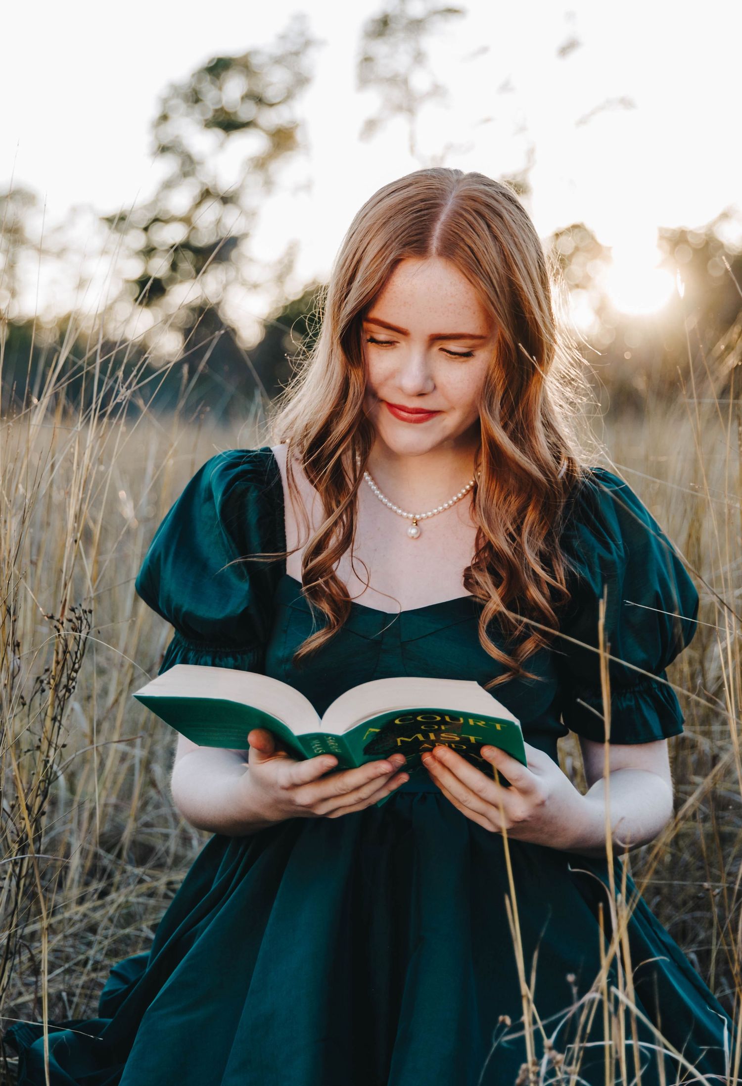 Young woman in emerald green dress reading a book in a golden field at sunset.