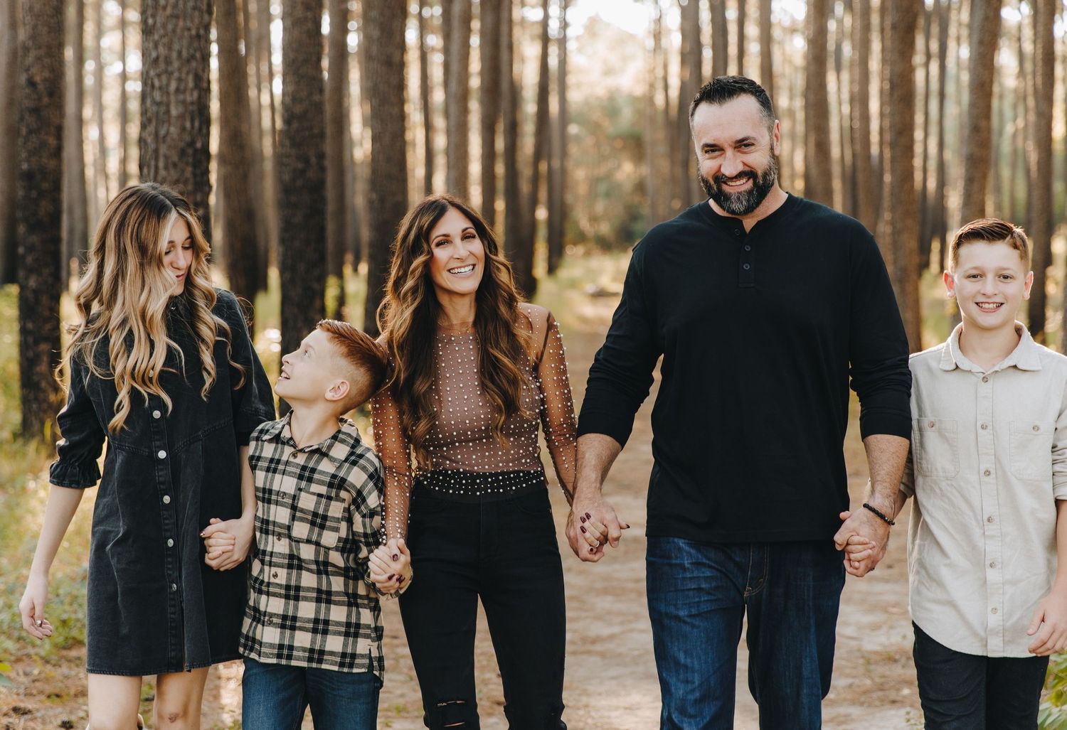 A family of five walks hand-in-hand smiling along a pine forest path during a family portrait session.