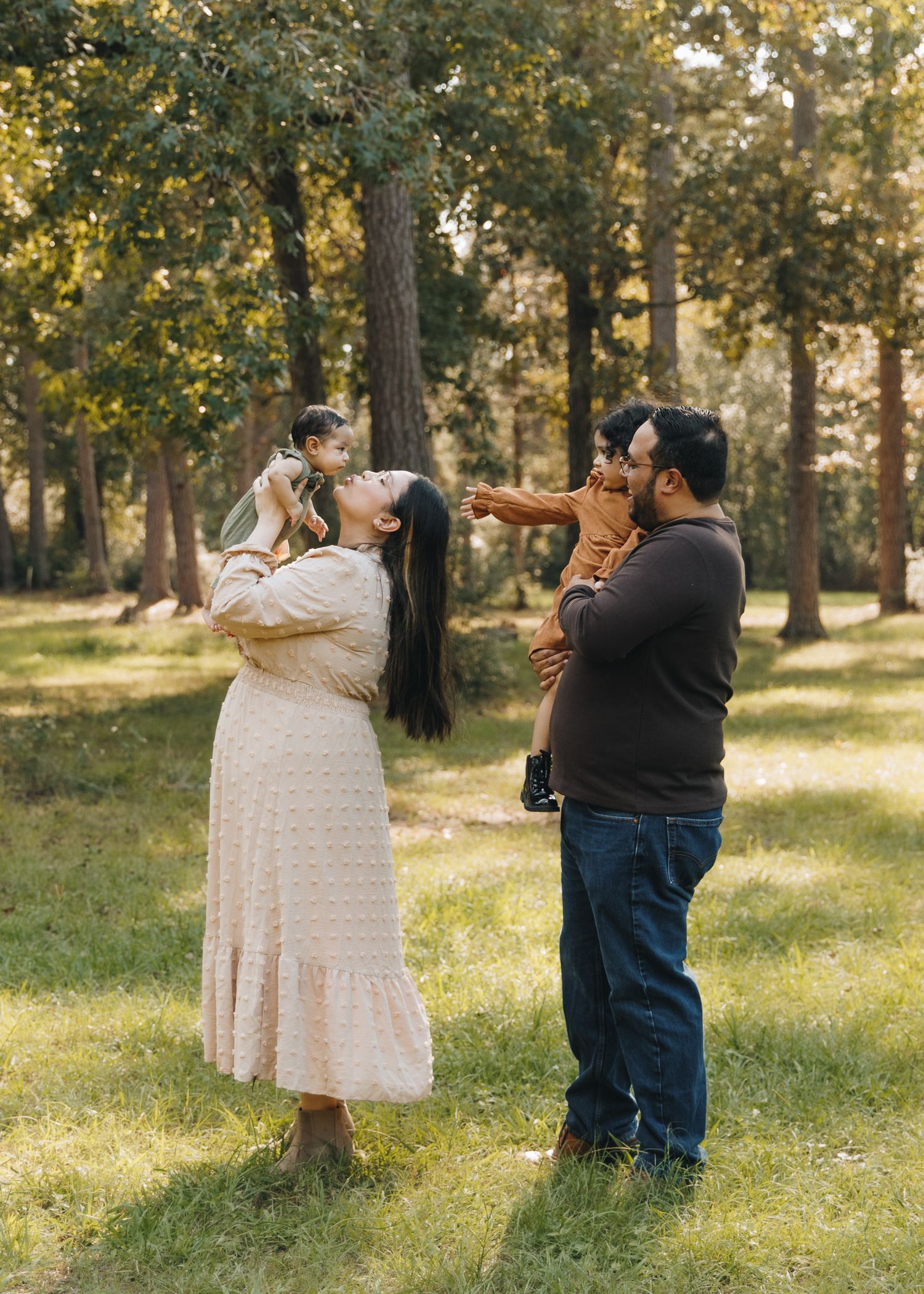 Family of four enjoying a sunny outdoor photo session in a green park, parents lifting two young children.