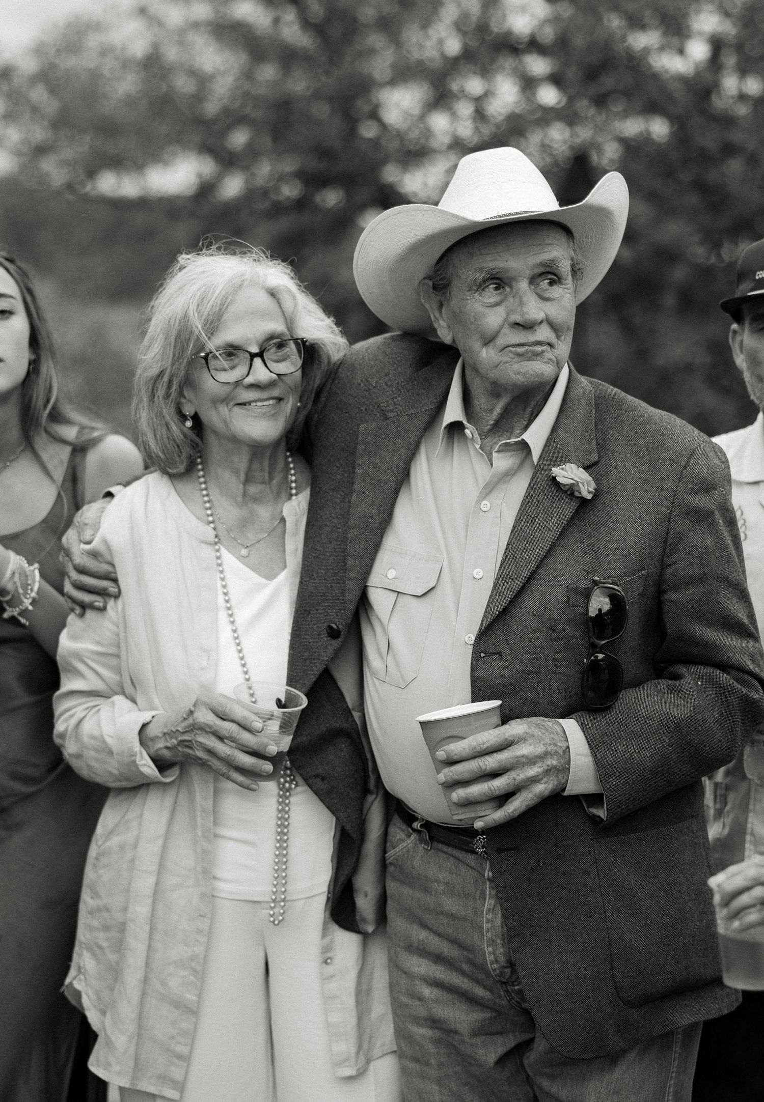 Elegant older couple smiling at an outdoor wedding, man wearing a white cowboy hat and blazer, woman in white dress with pearls.