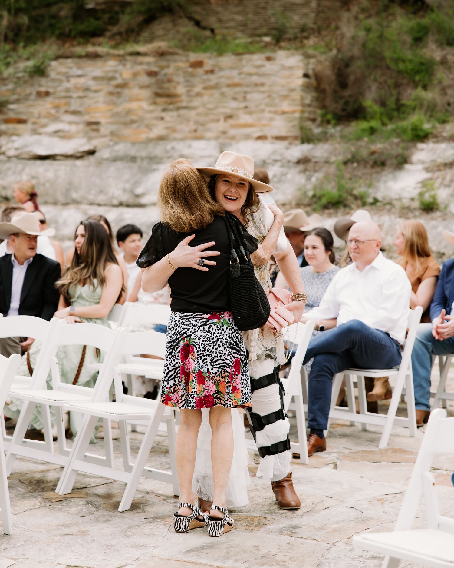 Two women share a warm hug in the aisle at an outdoor wedding ceremony with white chairs.
