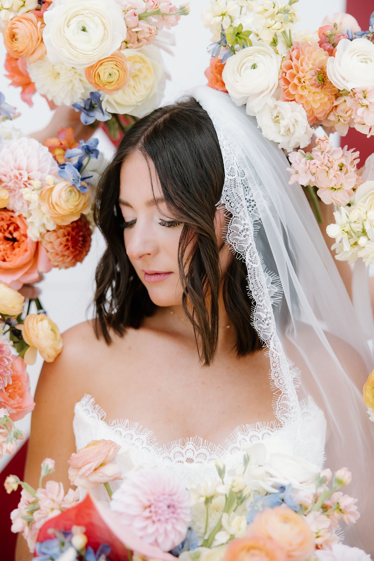 Beautiful bride in lace wedding dress and veil surrounded by colorful floral arrangements with soft gaze.