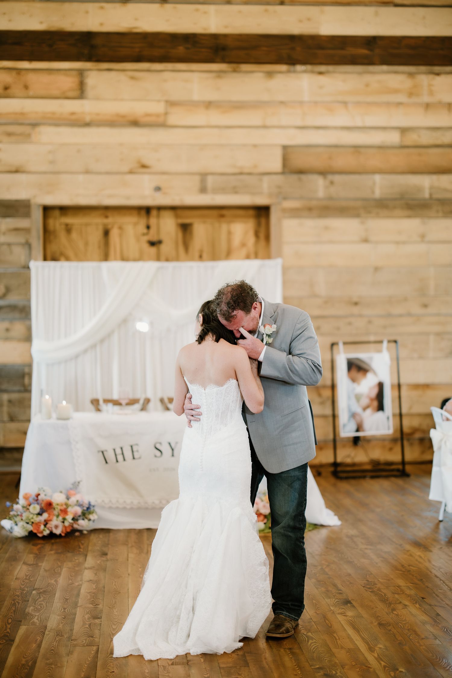 Bride and groom share an emotional first dance in rustic barn wedding venue with draped backdrop.