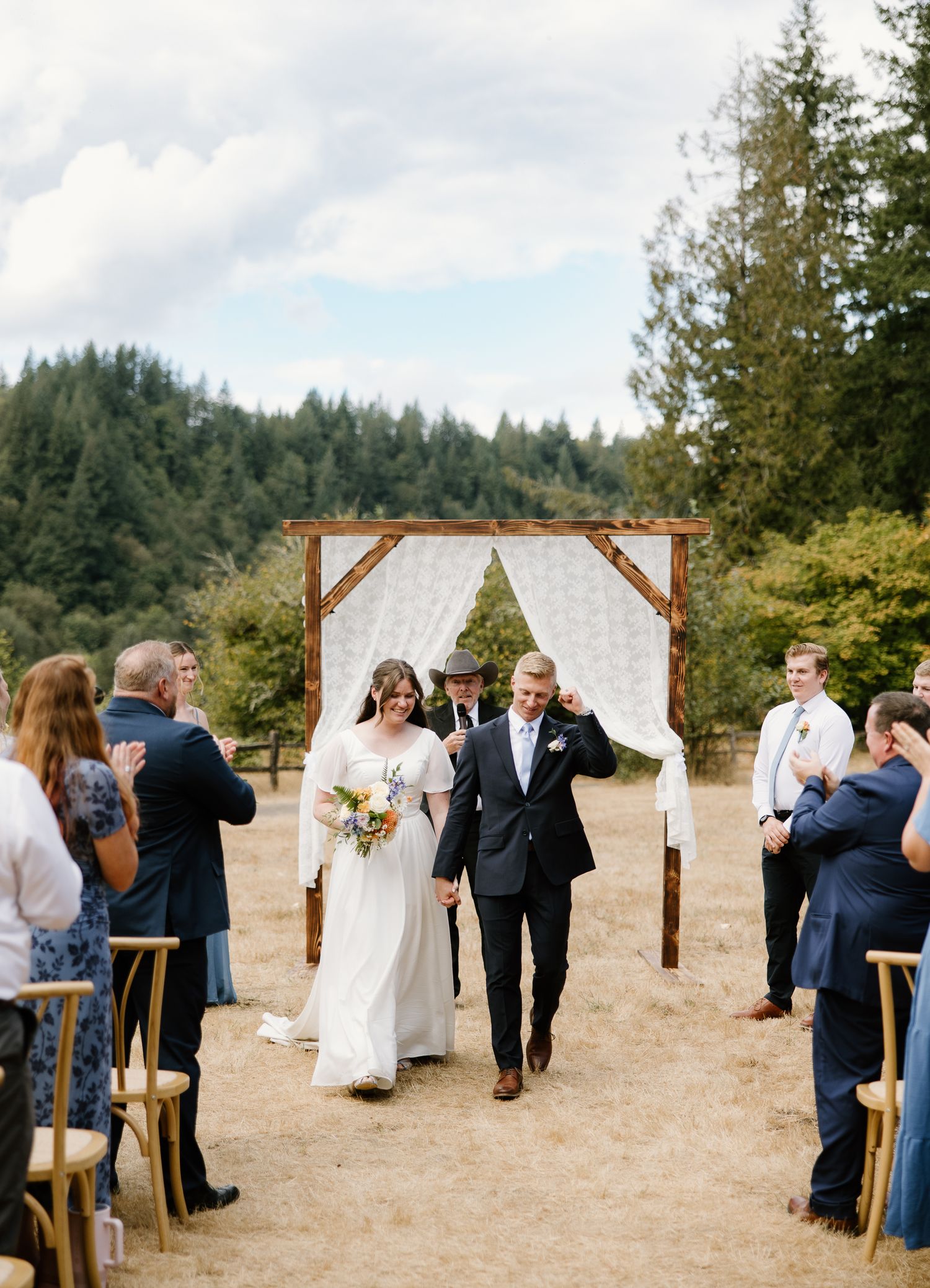 Newlyweds walk back down aisle after outdoor ceremony, guests applauding beside rustic wooden arch.